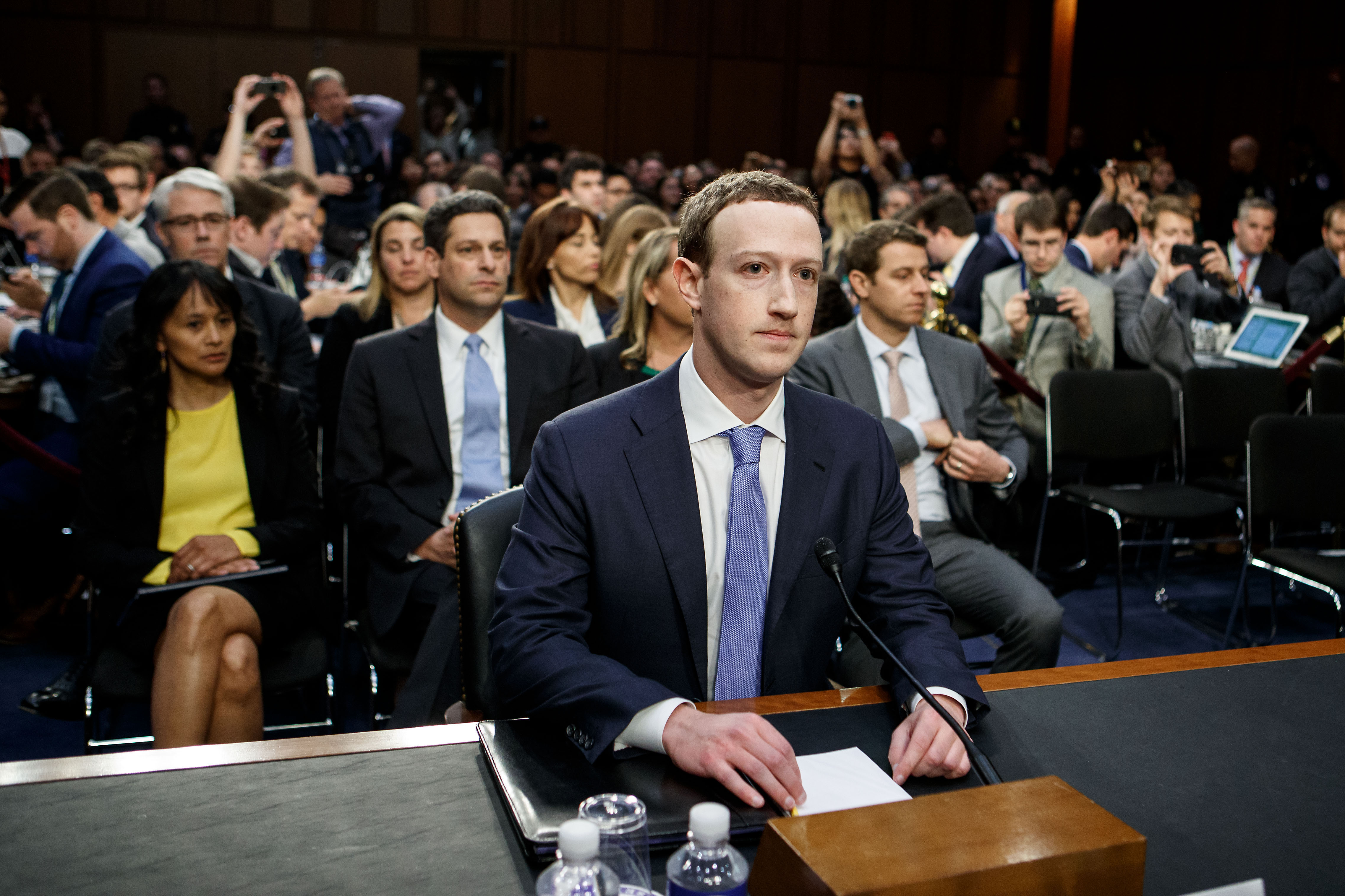 Facebook CEO Mark Zuckerberg sitting at a desk and wearing a suit and tie for his testimony before Congress in 2018. The room behind him is packed with seated people.