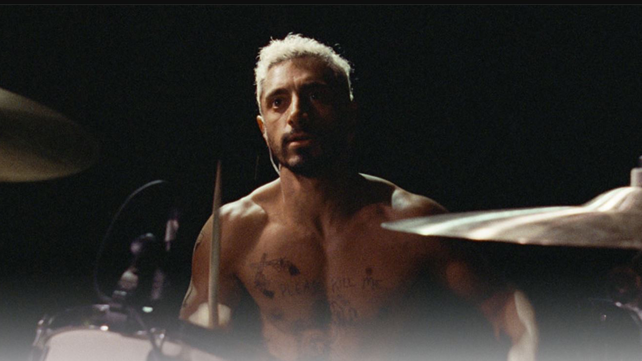 Ahmed, as Ruben, sits at a drum set shirtless against a black background.