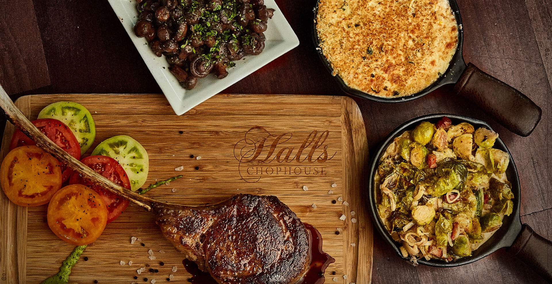 a cutting board labeled Halls Chophouse holds a large cut of meat, surrounded on the right by assorted side dishes