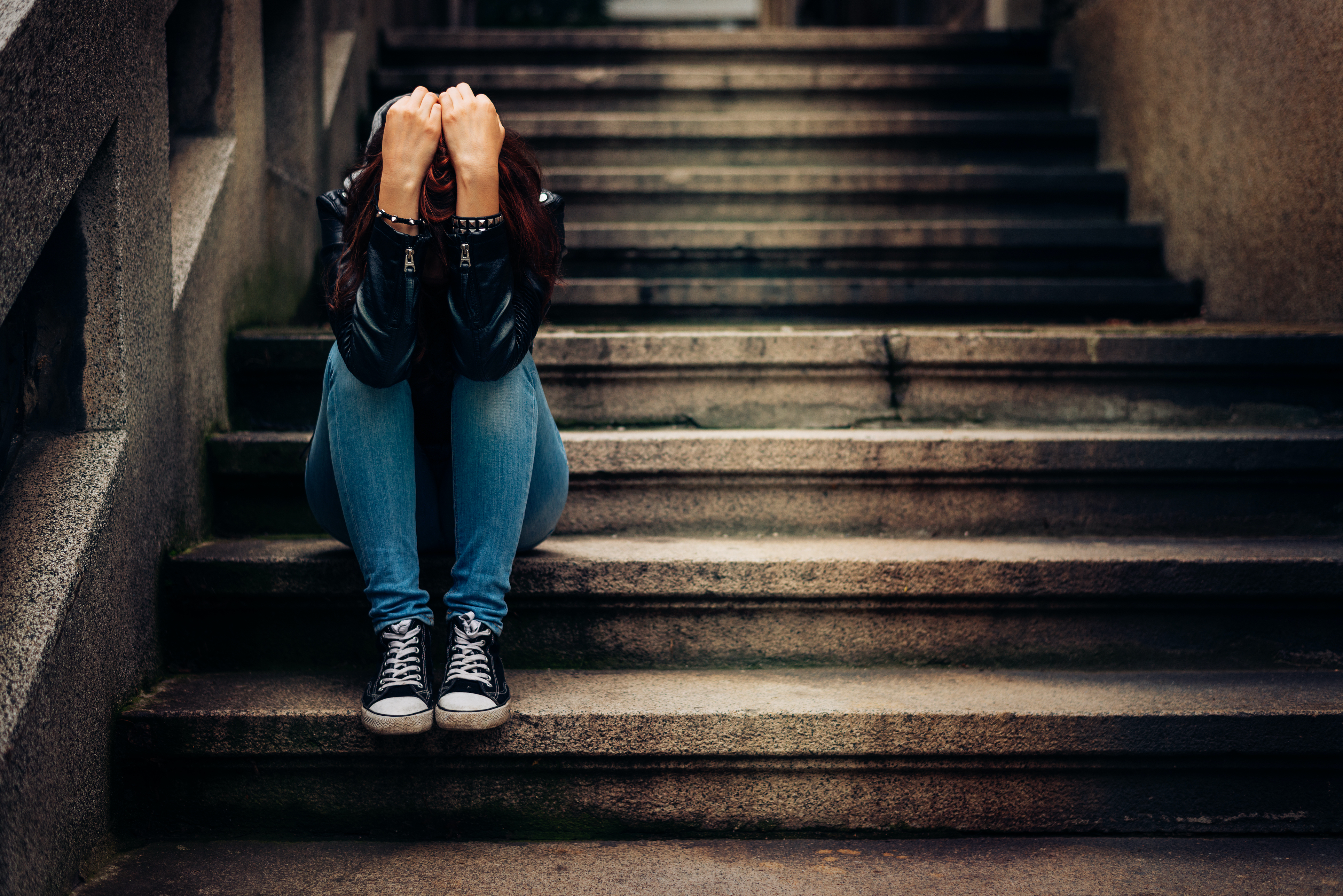 A young person wearing jeans and Converse-style shoes sits on concrete steps holding their head in their hands