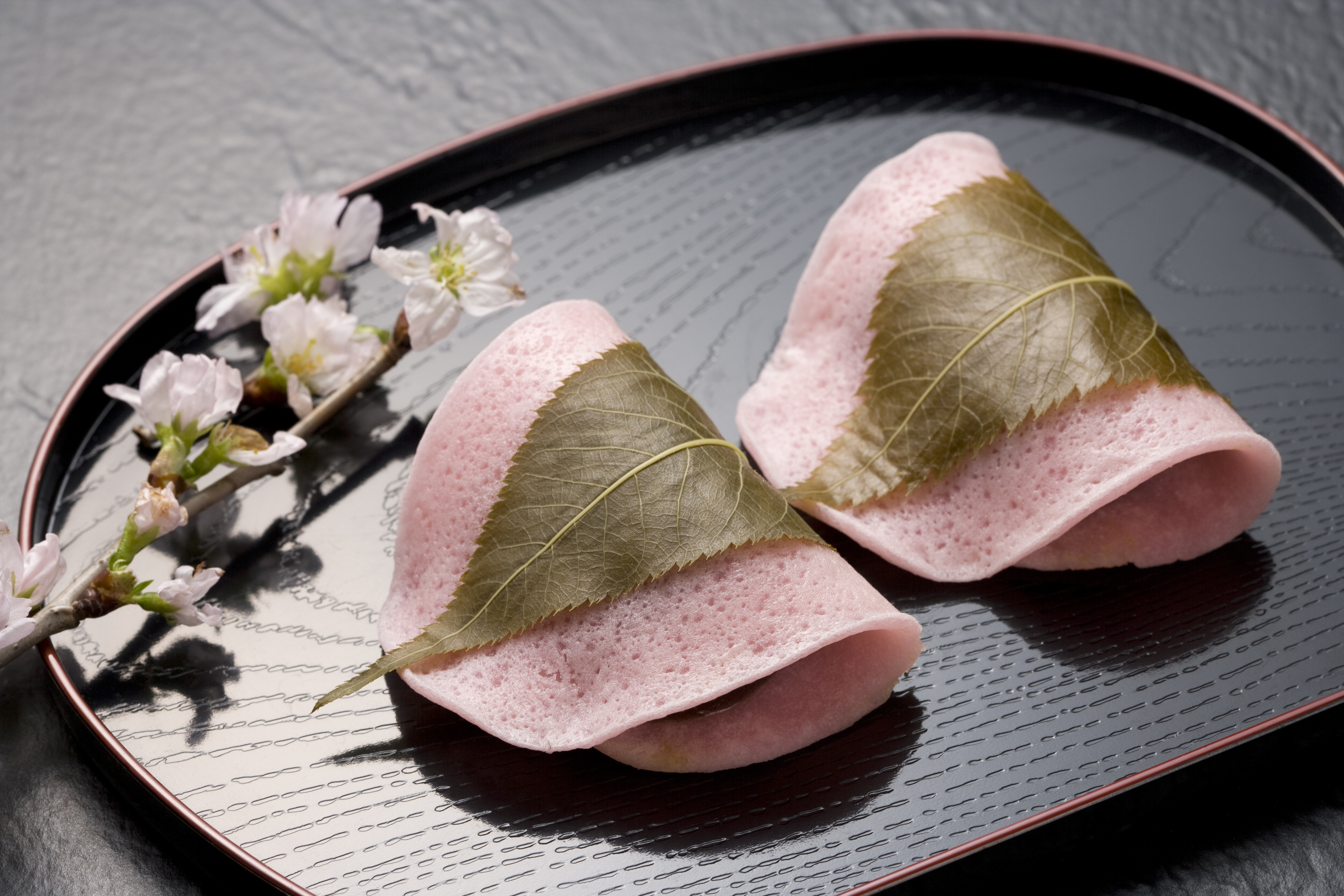 Two Sakura mochi — pink-hued rice cakes filled with red bean paste and wrapped in pickled cherry blossom leaves — on a black plate.