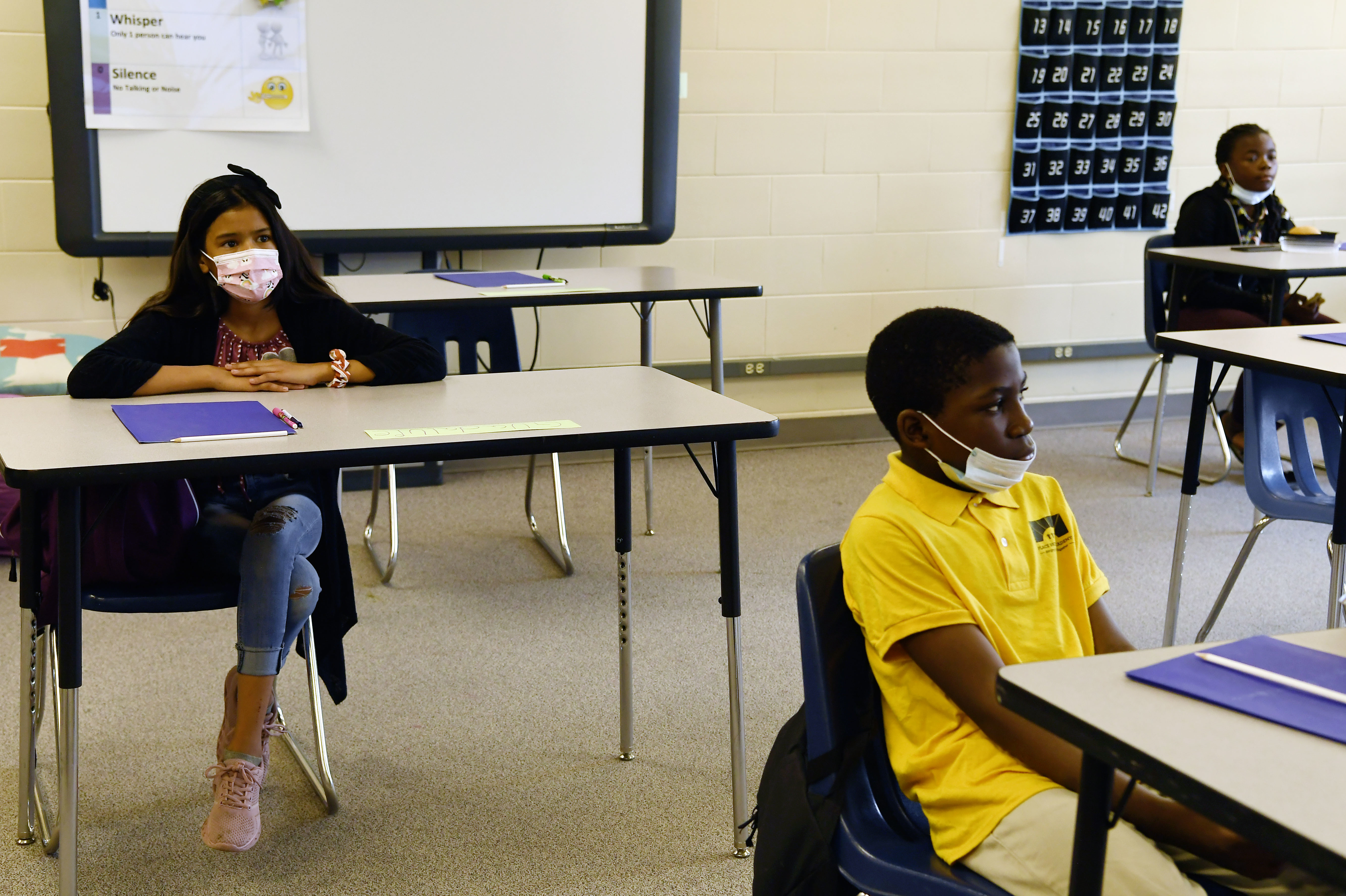 Three middle school students with masks sit at spaced out desks in a classroom, with a white board behind them.
