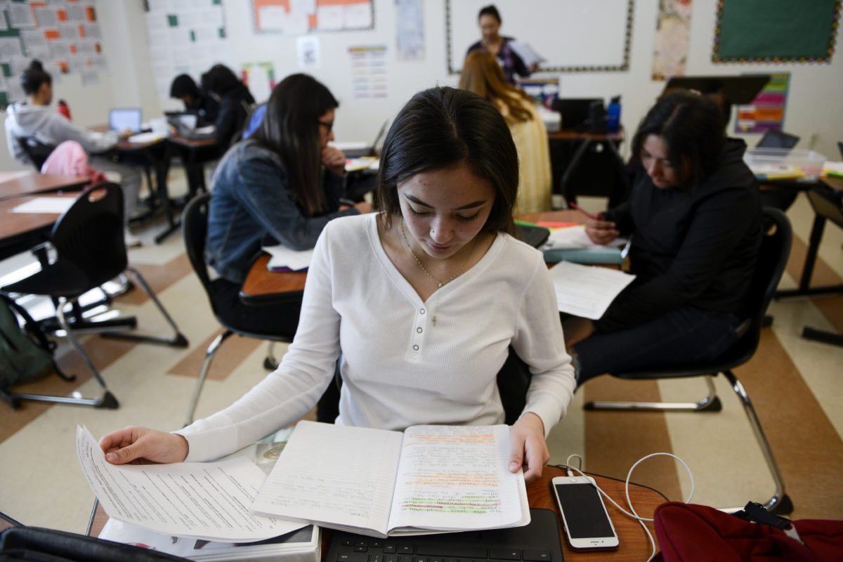 Adams City High School junior Alexandra Hernandez, in a white long-sleeve shirt, looks at her notebook, with a cell phone next to her. In the background are about five students working at tables and a teacher standing in front of a white board, in an AP language class  on Feb. 4, 2019.