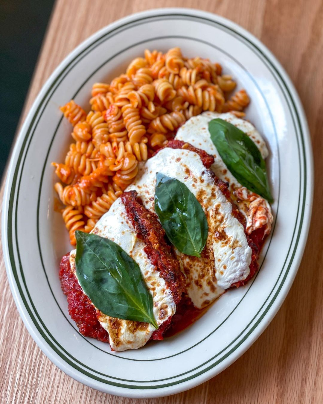 Overhead view of chicken parm on a white plate with a side of rotini. The plate is on a light wooden surface.