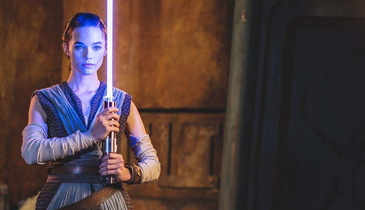 An actress playing Rey holds a blue prop lightsaber in vertical position.