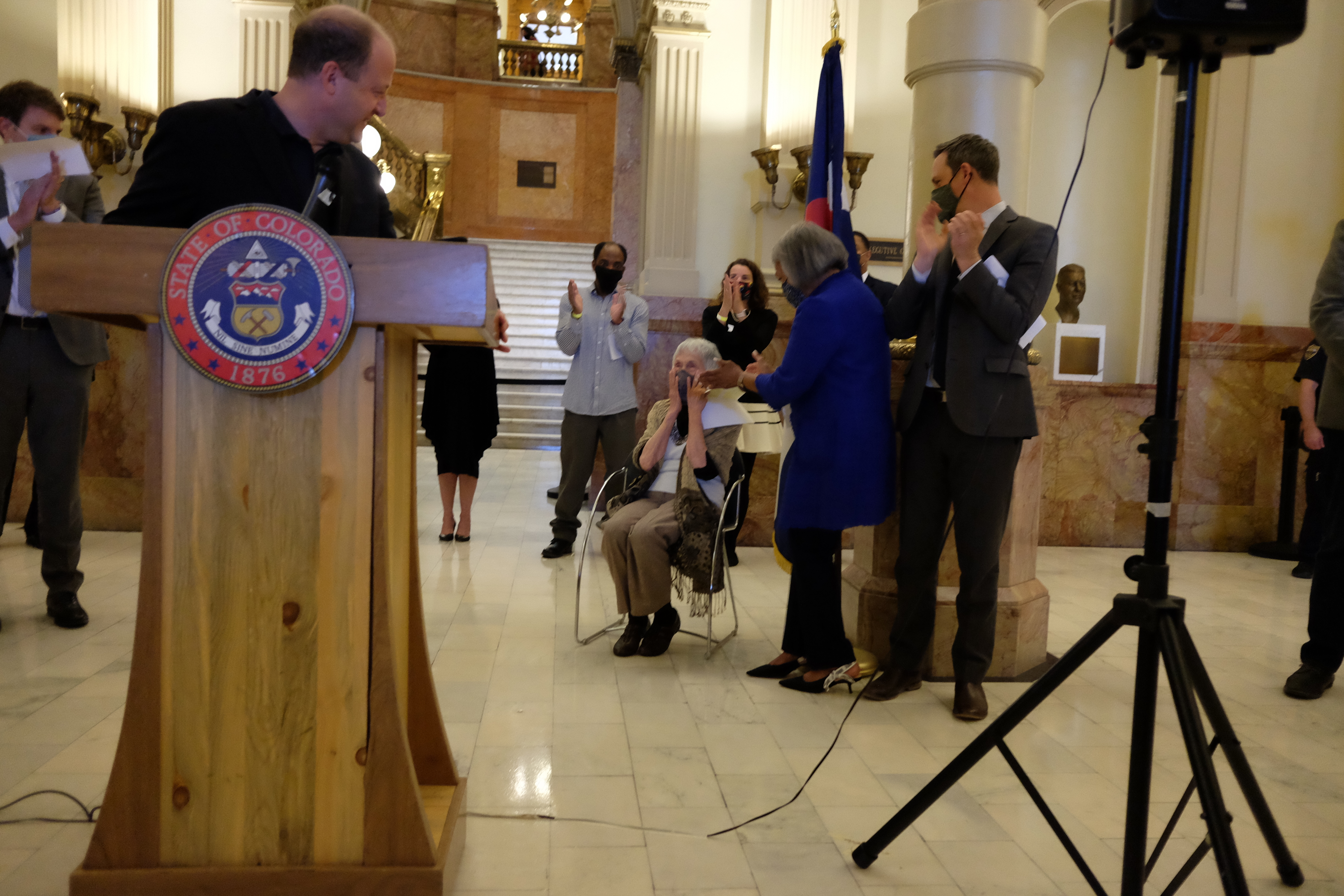 Gov. Jared Polis stands at a podium and turns to look at early childhood advocate Anna Jo Haynes, seated. She clasps her hands to her face in surprise. Lawmakers standing nearby applaud.