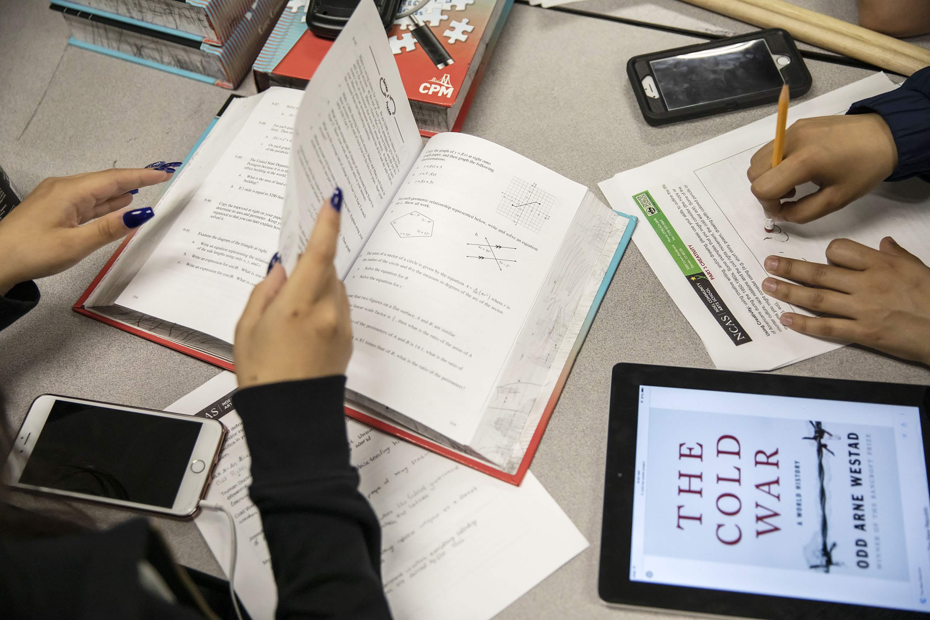 Hands of a student with black fingernail polish turn the pages of a math book, with an iPhone nearby. At the same table, the hands of another student erase a mark on a worksheet, with a tablet device depicting a book or lesson, “The Cold War,” nearby.