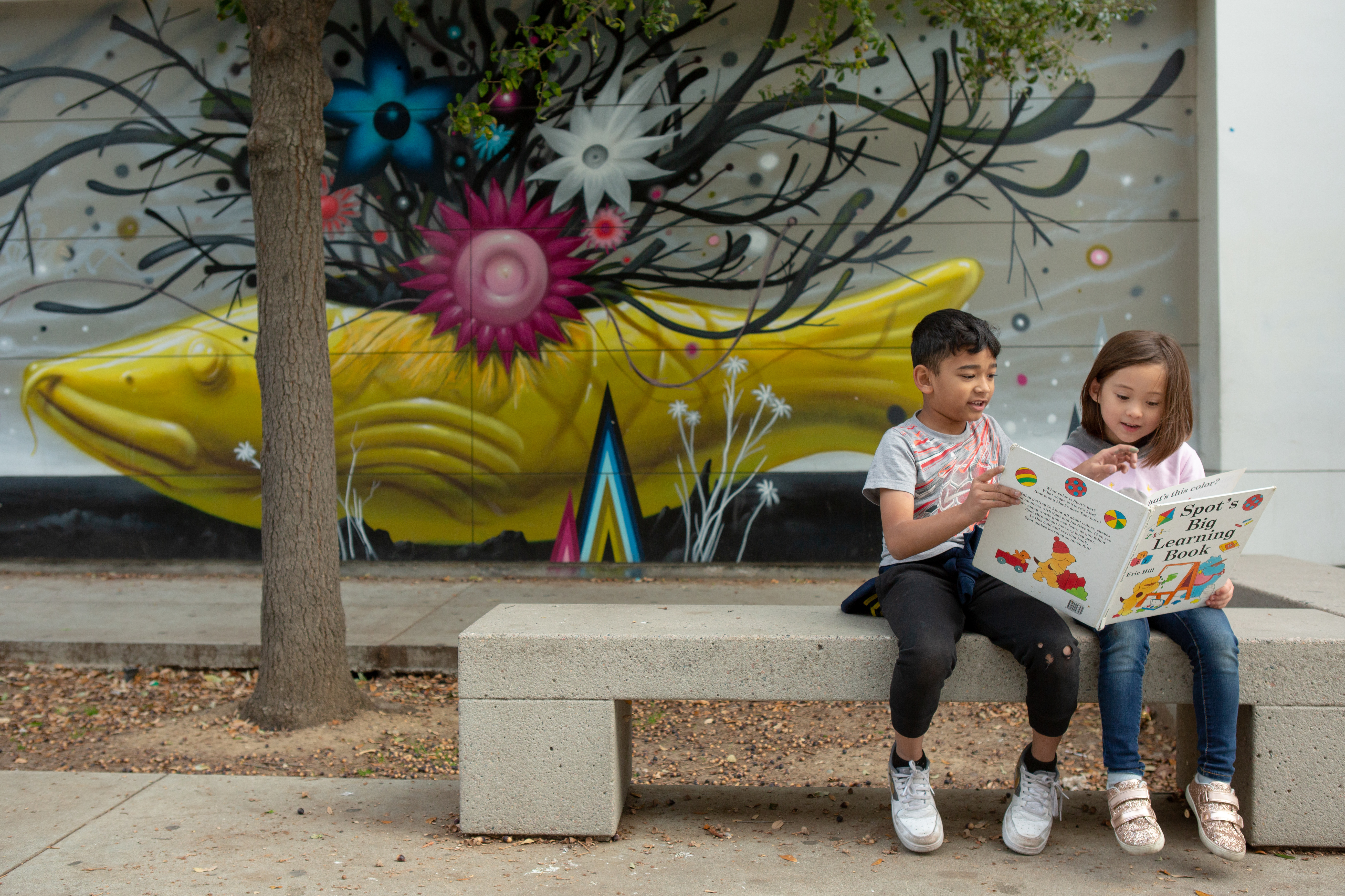 A boy and a girl share a picture book while sitting on a concrete bench in front of a concrete wall and a sculpture of a large yellow fish with a tangle of dark seaweed growing up from its back with blue, magenta and white flowers interspersed.