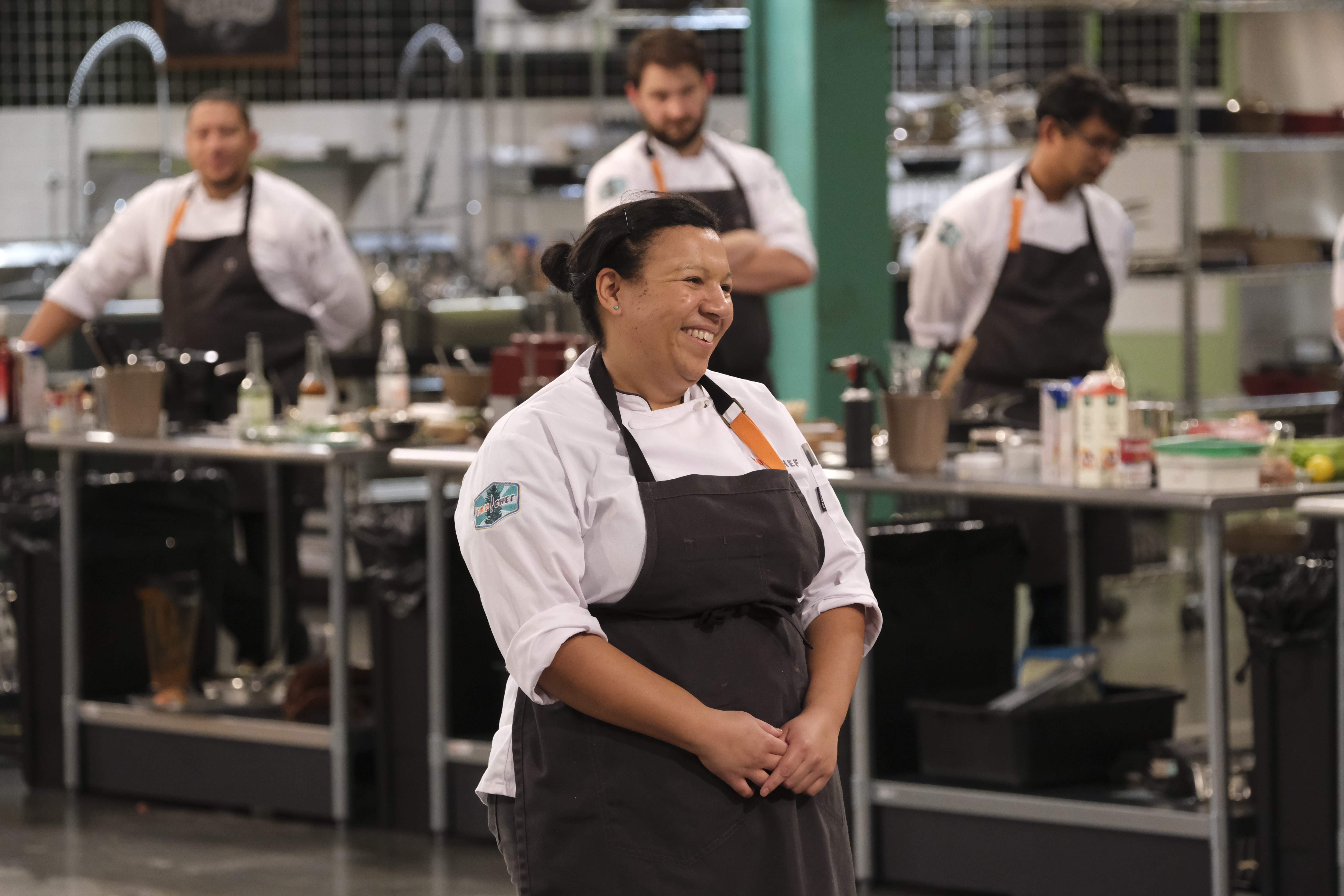 A smiling woman with an apron stands in the foreground of a large room. Three men with aprons are standing in the background.