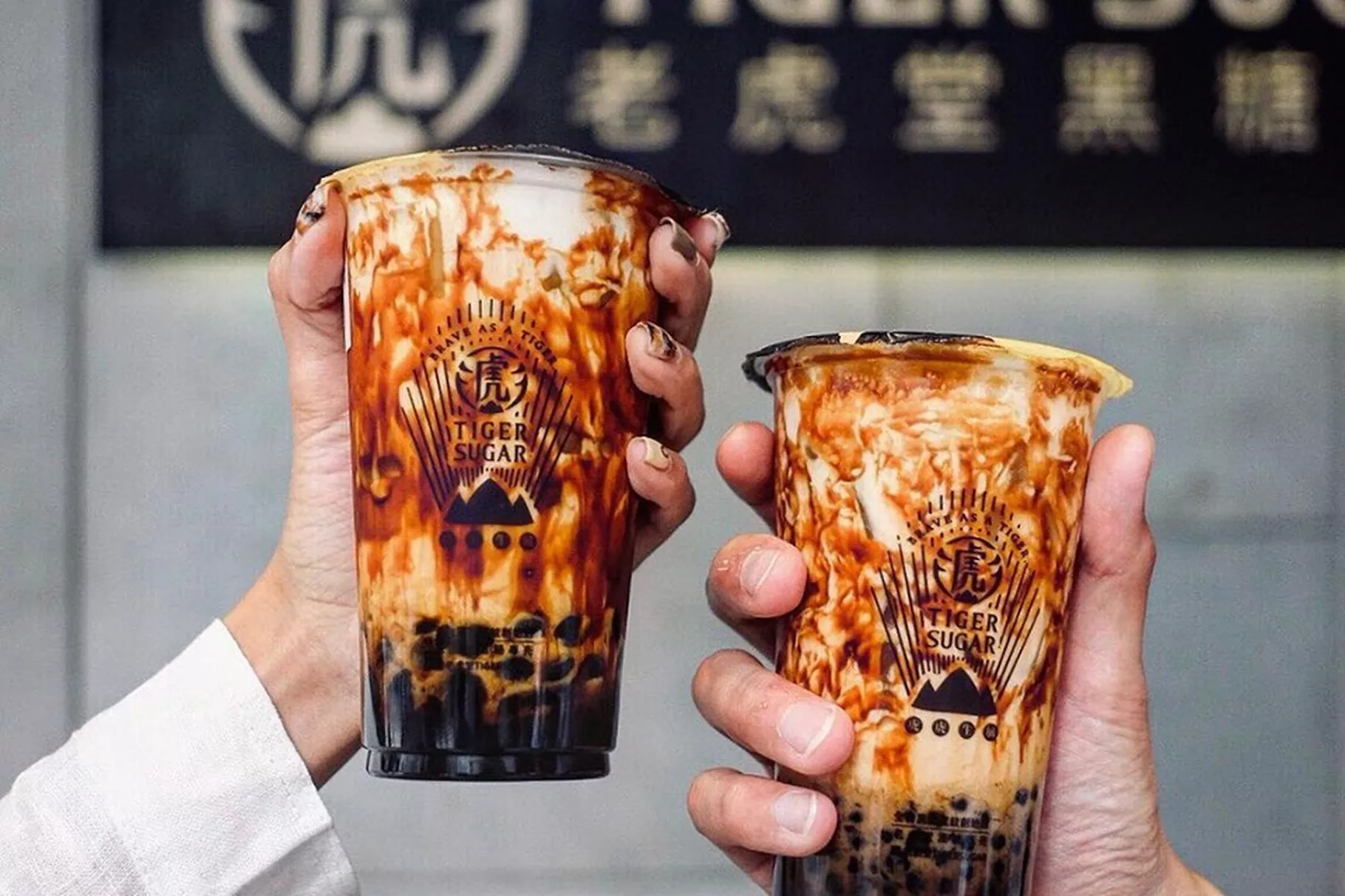 Two hands hold cups of brown sugar boba from Taiwanese chain Tiger Sugar. The iced tea drink is milky white with streaks of dark brown.