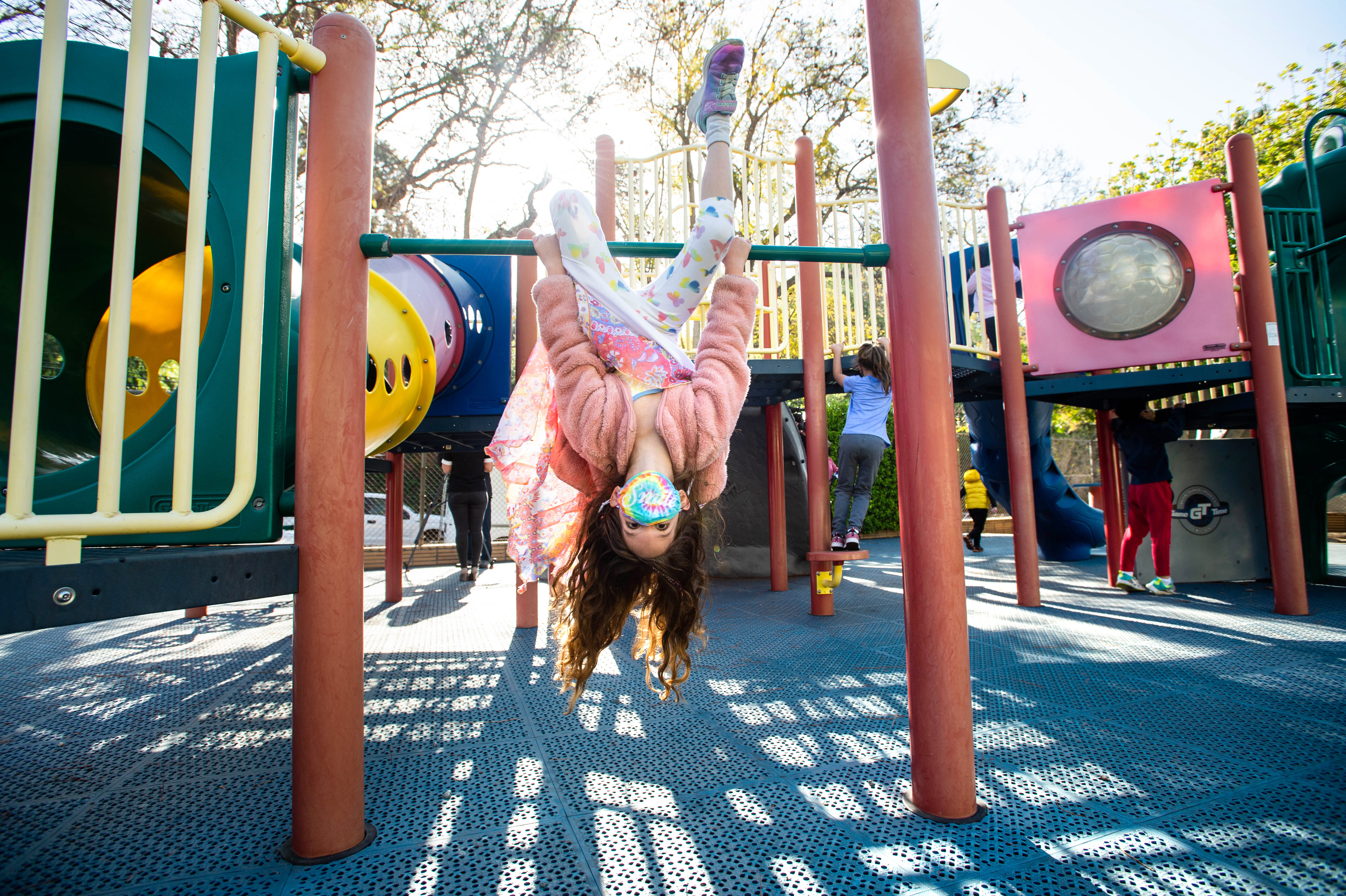 A masked kindergartner hangs upside down from playground equipment, her long hair dangling toward the ground.