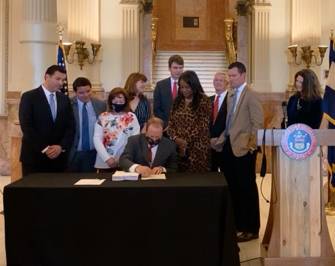 Seated at a table covered by a dark cloth, Gov. Jared Polis signs a piece of paper next to a large stack of documents. Colorado legislators stand around him looking on.
