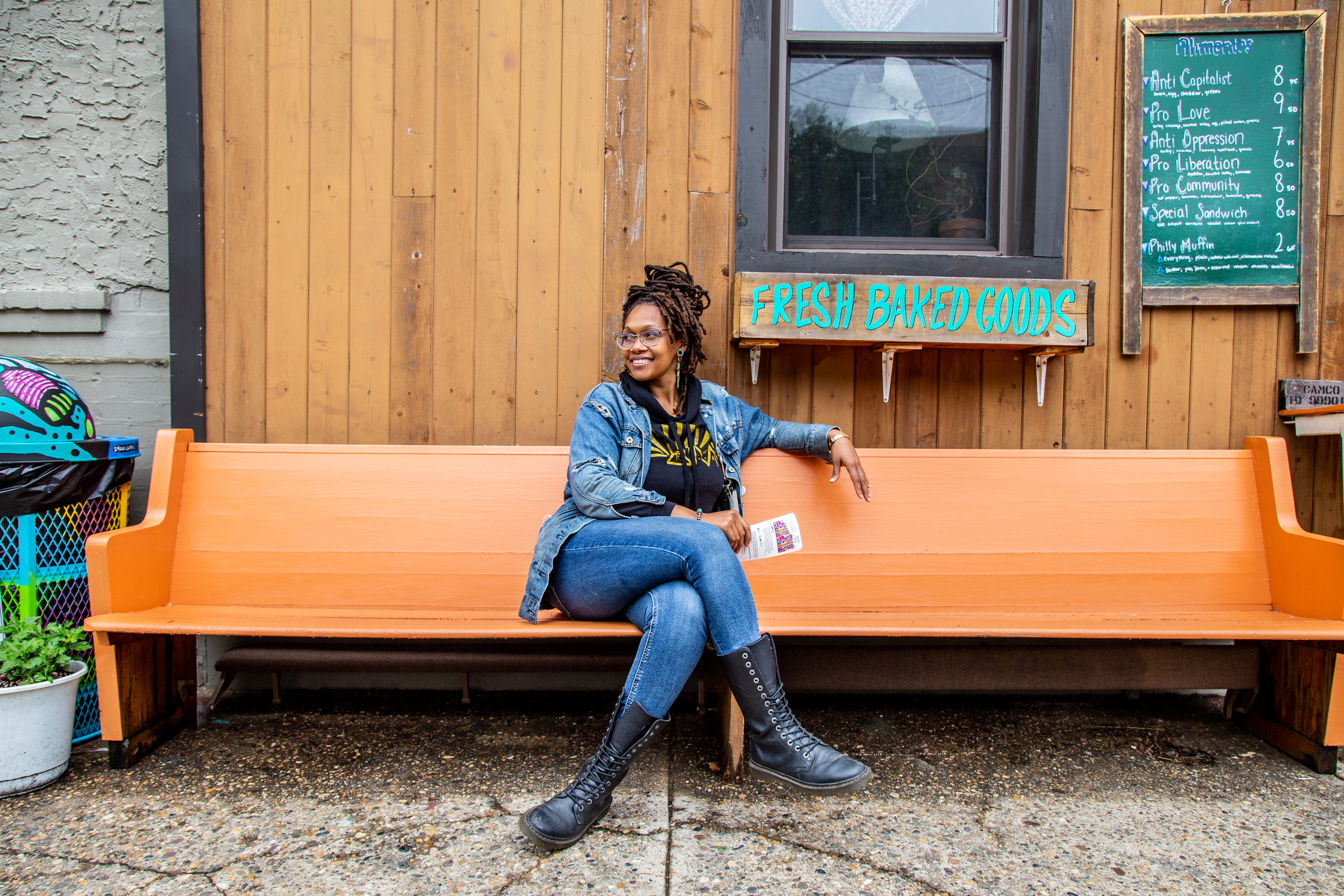 Founder of Franny Lou’s Porch, Blew King, sits cross-legged in jeans and black boots on an orange bench at a coffee shop