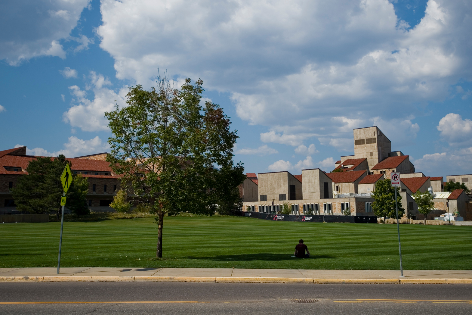 A University of Colorado Boulder student studies in the shade of a tree in front of Business Field with school buildings in the background.