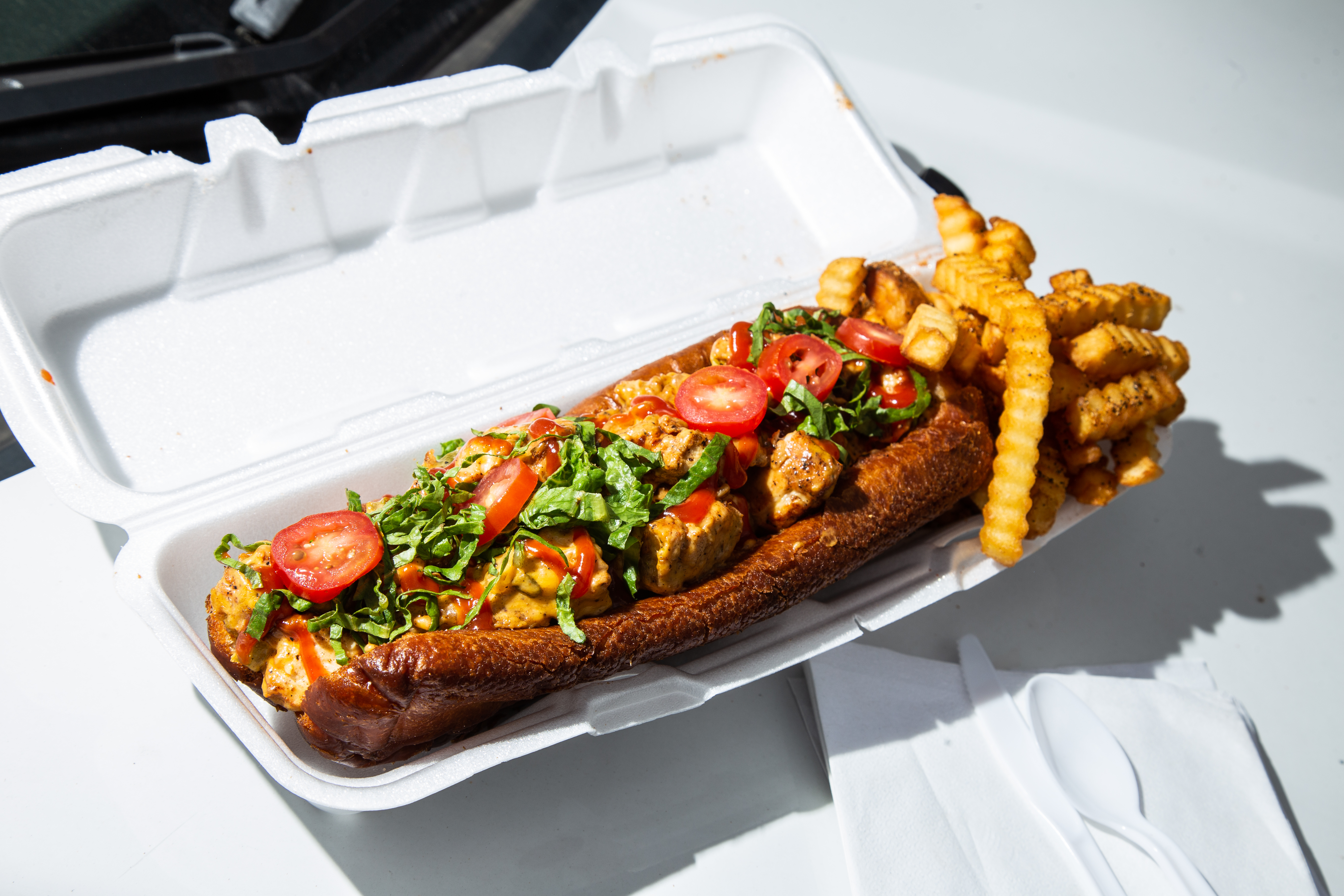 a deep fried chicken cheesesteak with cherry tomatoes, lettuce, special sauce, and crinkle fries in a styrofaum container