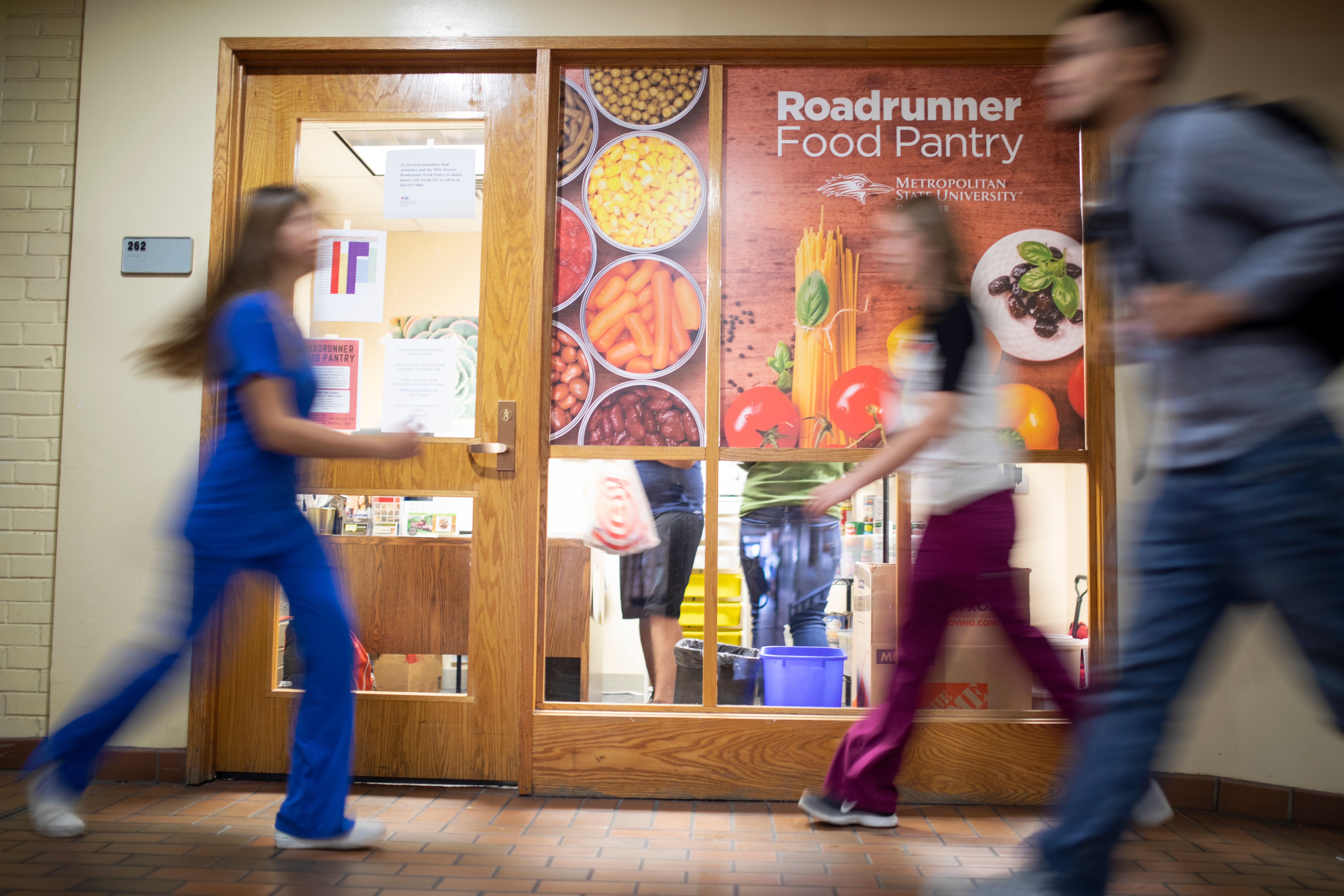 Blurred students walk past a Metropolitan State University of Denver door with a colorful sign that reads: Roadrunner Food Pantry.