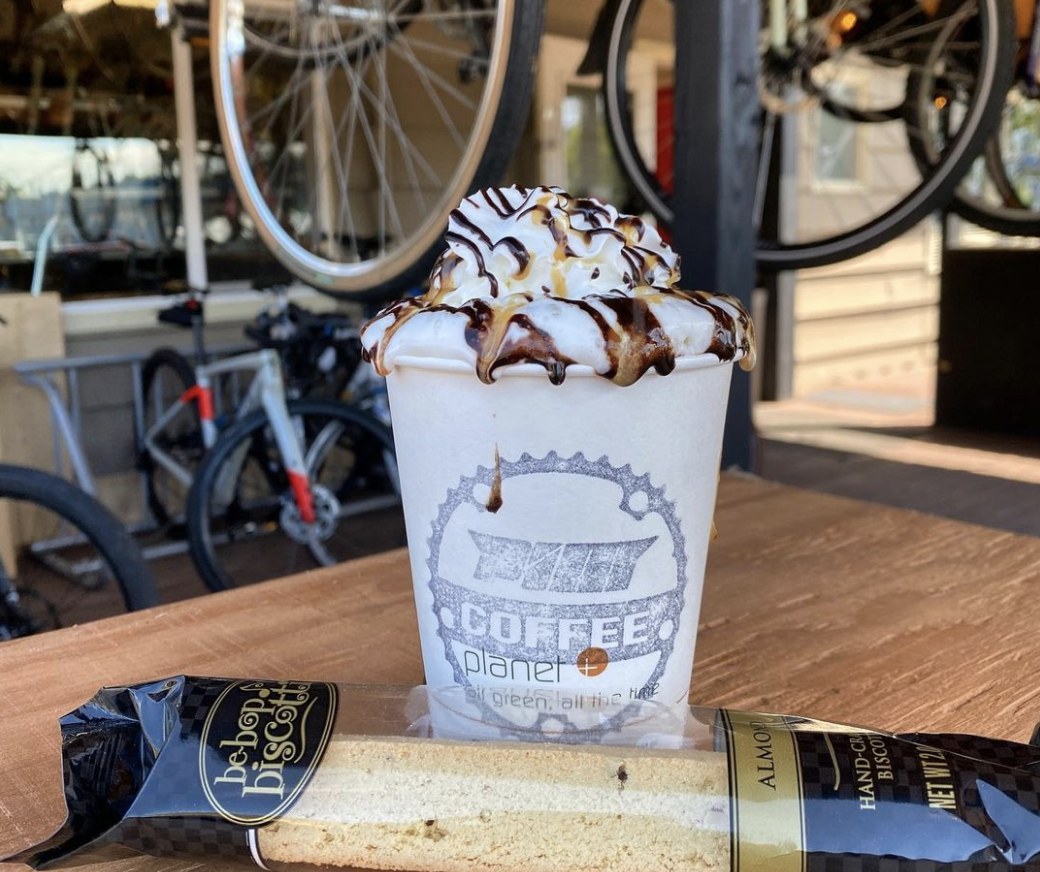 A whipped cream-topped coffee drink in a paper cup next to a wrapped biscotti, with bicycles at a shop hanging in the background