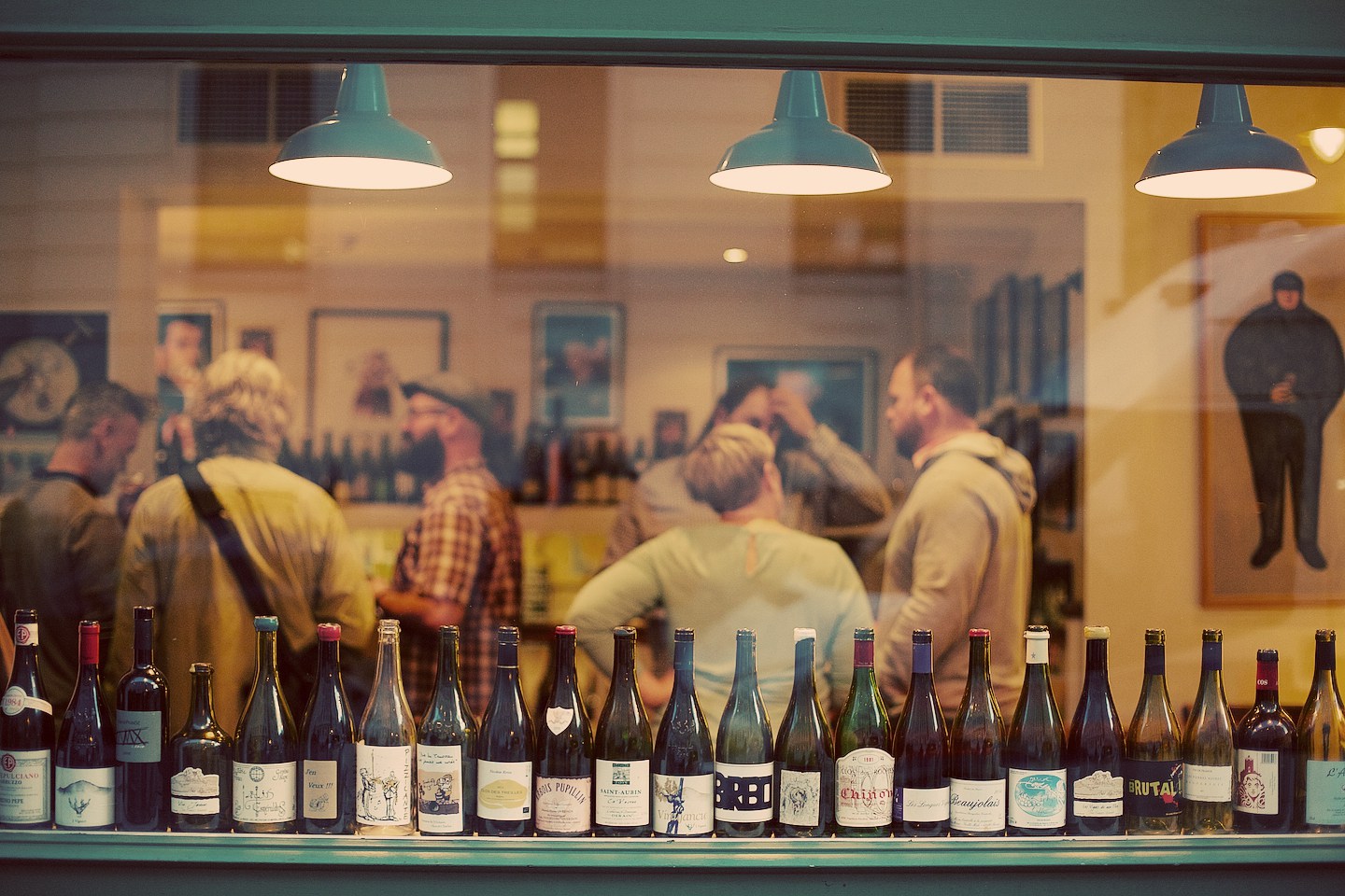 A crowd of wine drinkers seen through a front window, its sill lined with emptied bottles.