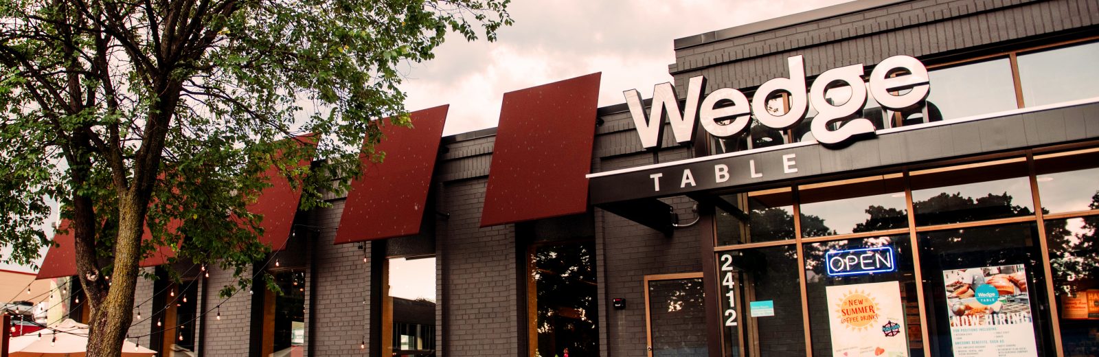 The exterior of The Wedge Table with a large white letters spelling out Wedge and the word table smaller underneath. The new-construction-style modern building has black/gray fixtures and large panel glass windows