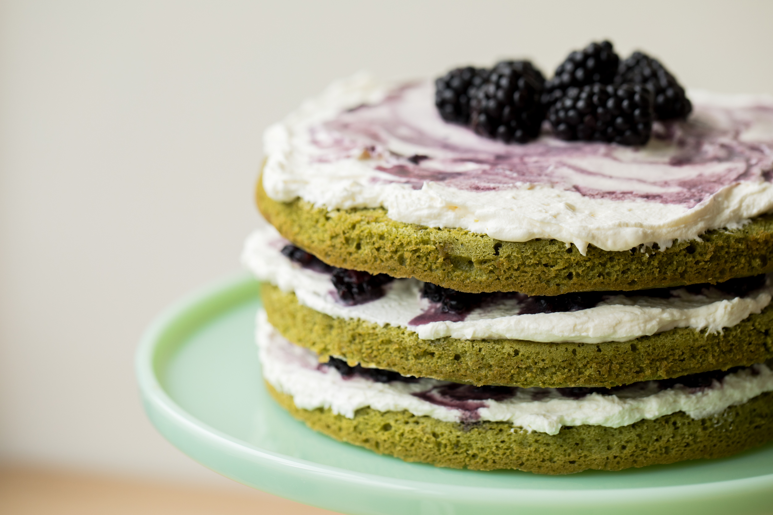 A side view of a matcha blackberry layer cake: the four green layers are sandwiched with whipped cream and mashed blackberries, and the top is covered with whipped cream and whole blackberries. The cake sits on a jade cake stand.