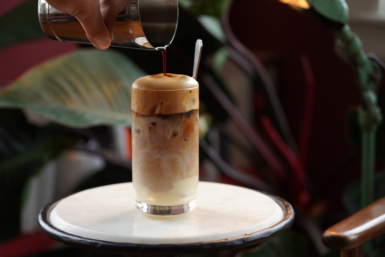 A hand pours liquid into a glass full of a foamy, milky iced coffee on a small white table. Plants are visible in the background.