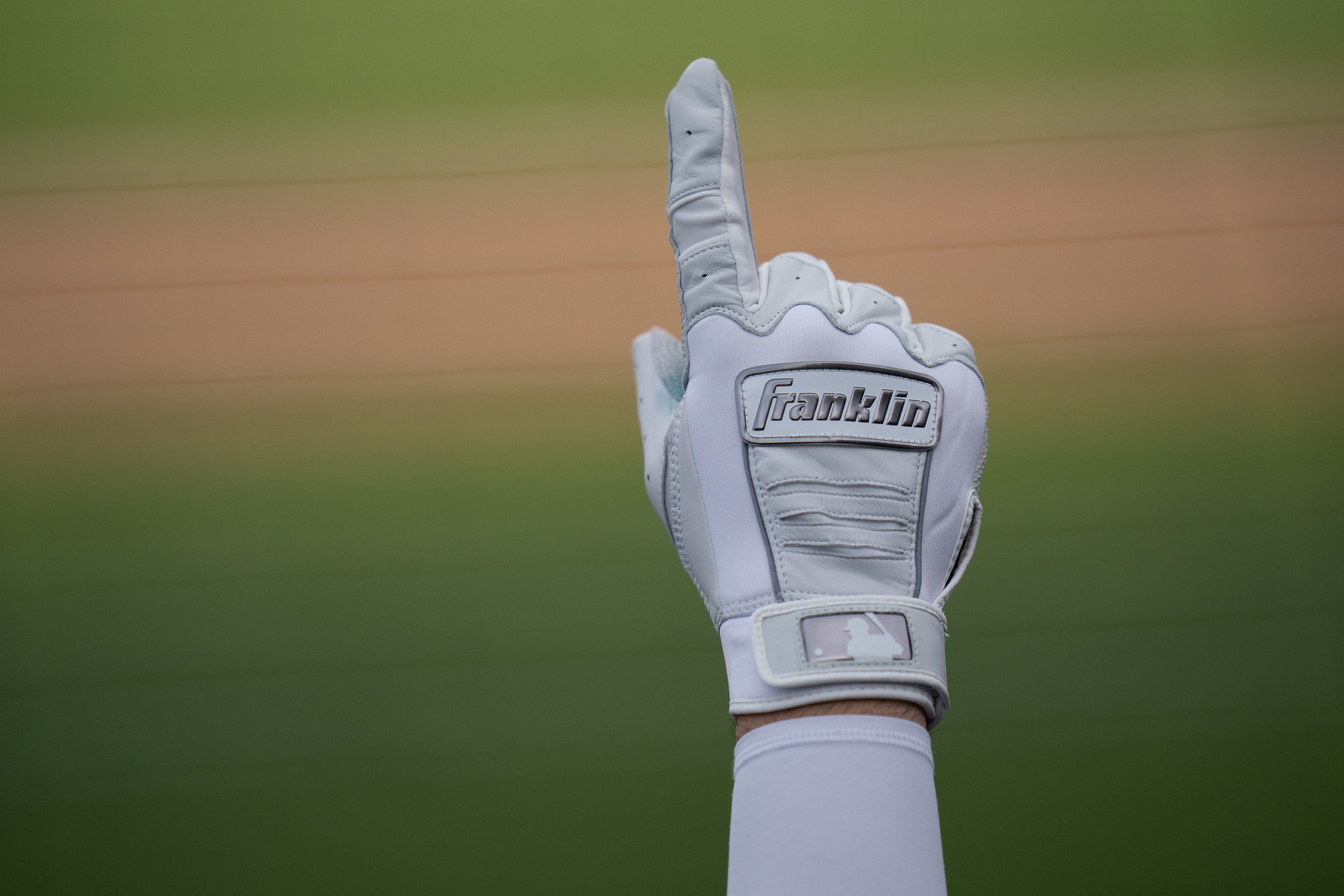 A hand clad in a white Franklin brand batting glove with silver accents points up in the air, and is the only thing visible against a distant backdrop of the green grass and brown dirt of a baseball diamond.