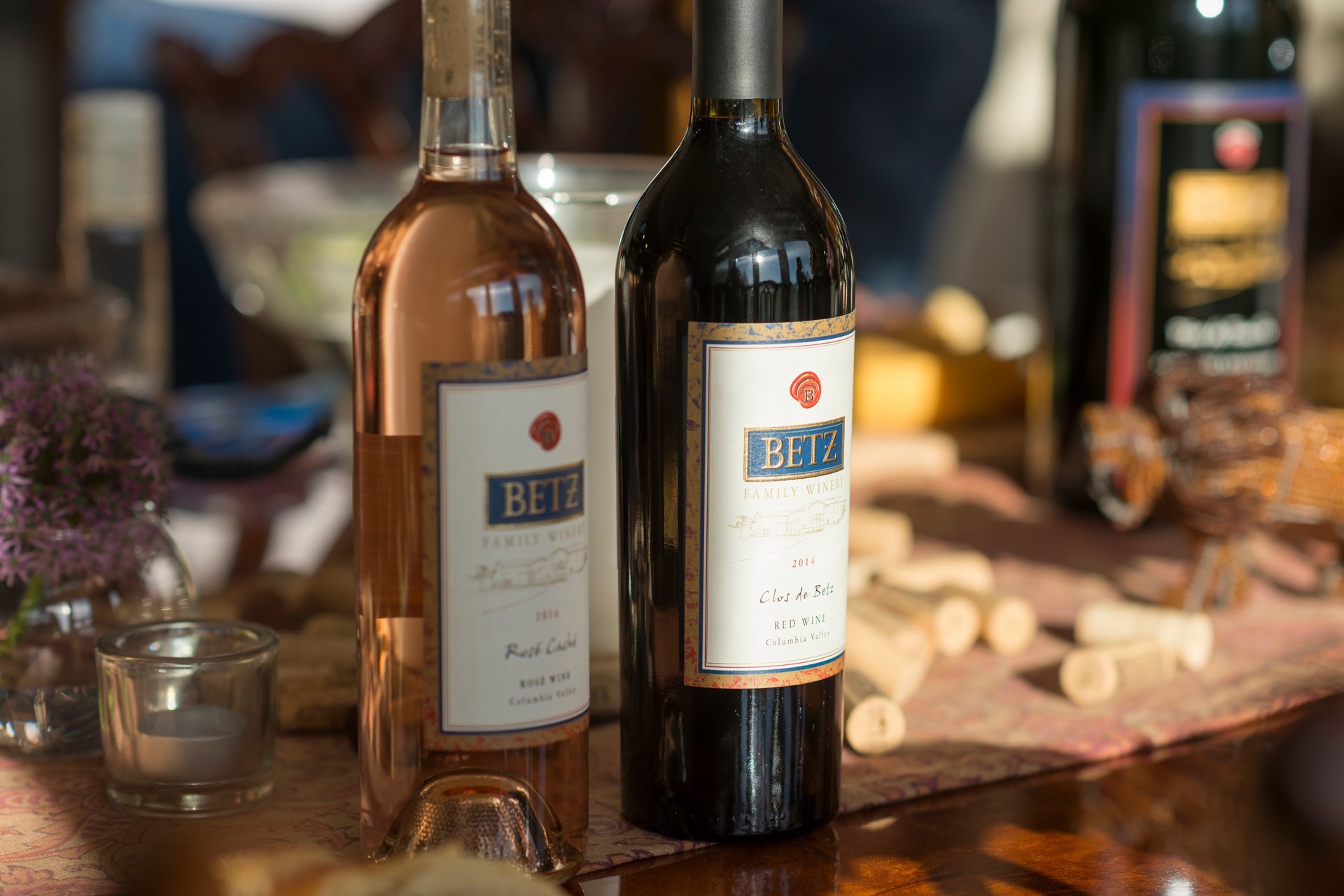 A bottle of red wine to the right; a bottle of white to the left with the Betz Family WInery label on both, and a cheeseboard in the backrgound