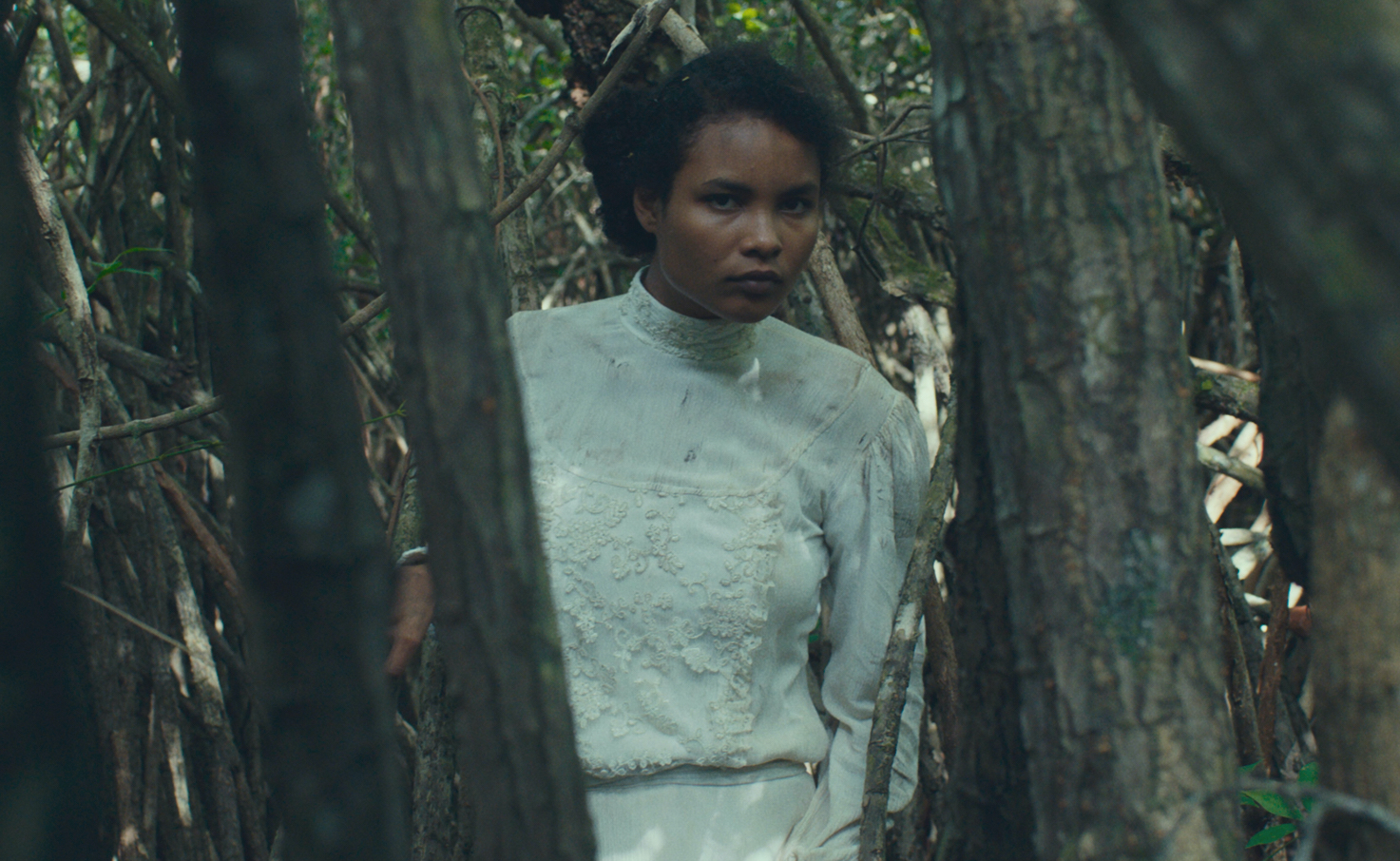 A woman in white, played by Indira Rubie Andrewin, peers between trees in Tragic Jungle