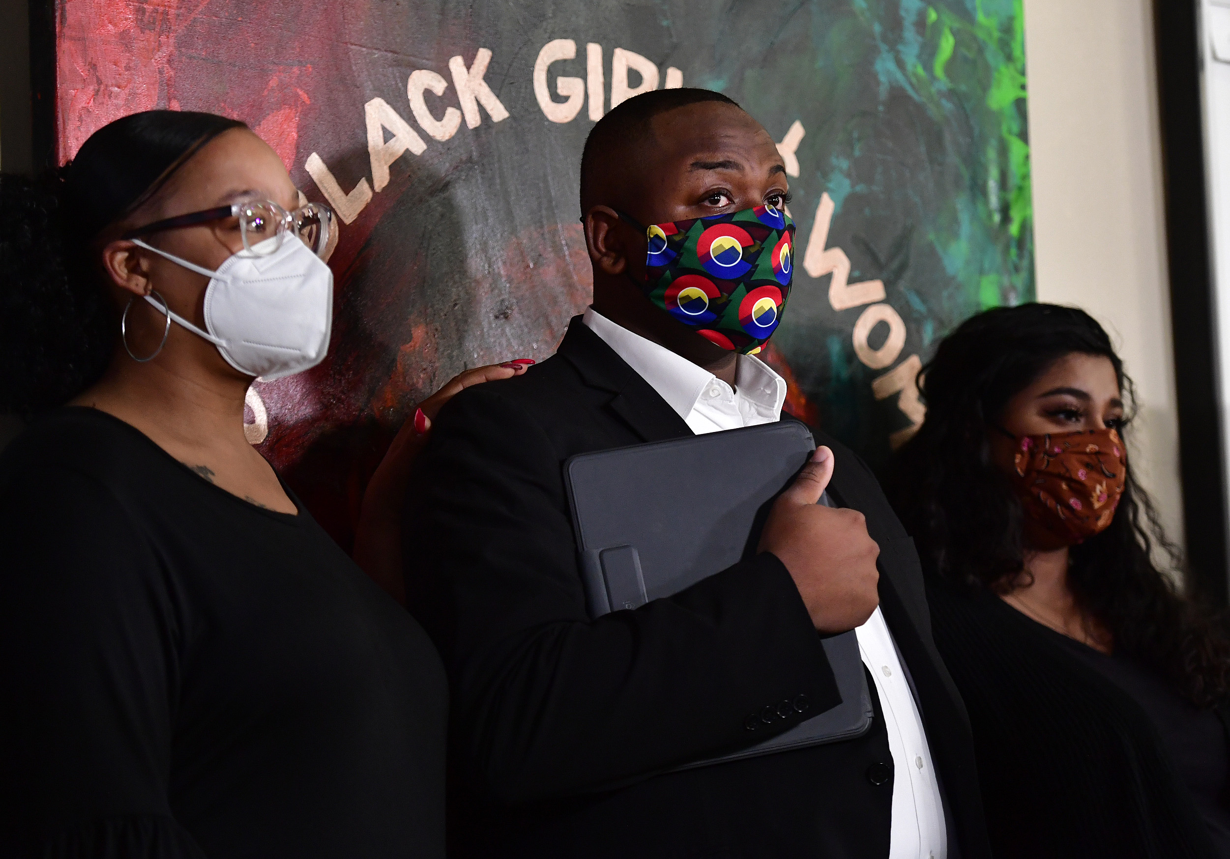 Tay Anderson, wearing a face mask, stands with two women, one on either side of him. A painting on the wall behind him reads “Black Girls and Women.”