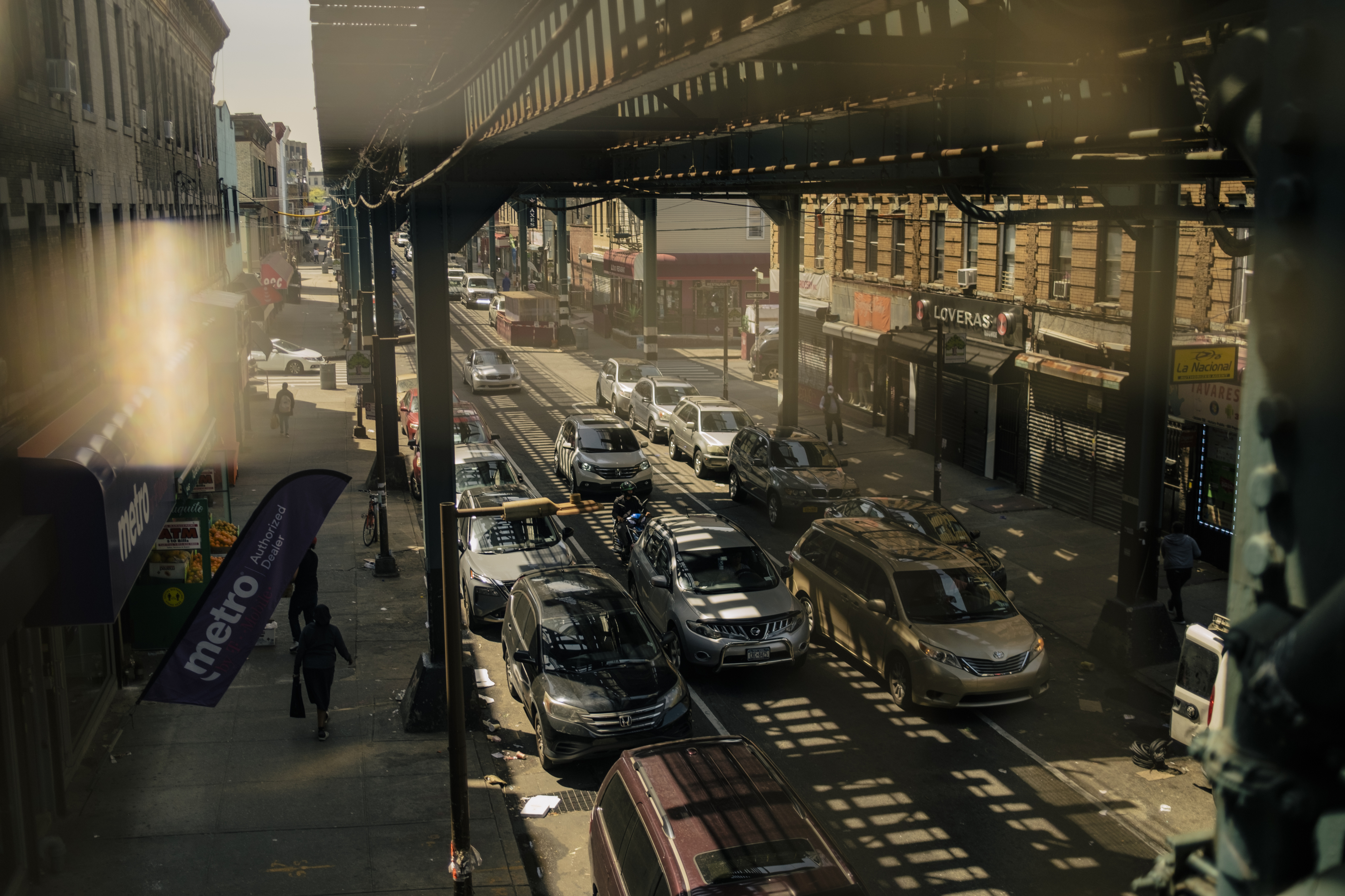 A bustling street in Cypress Hills, Brooklyn, with traffic moving under a subway track and shadows lining the street.