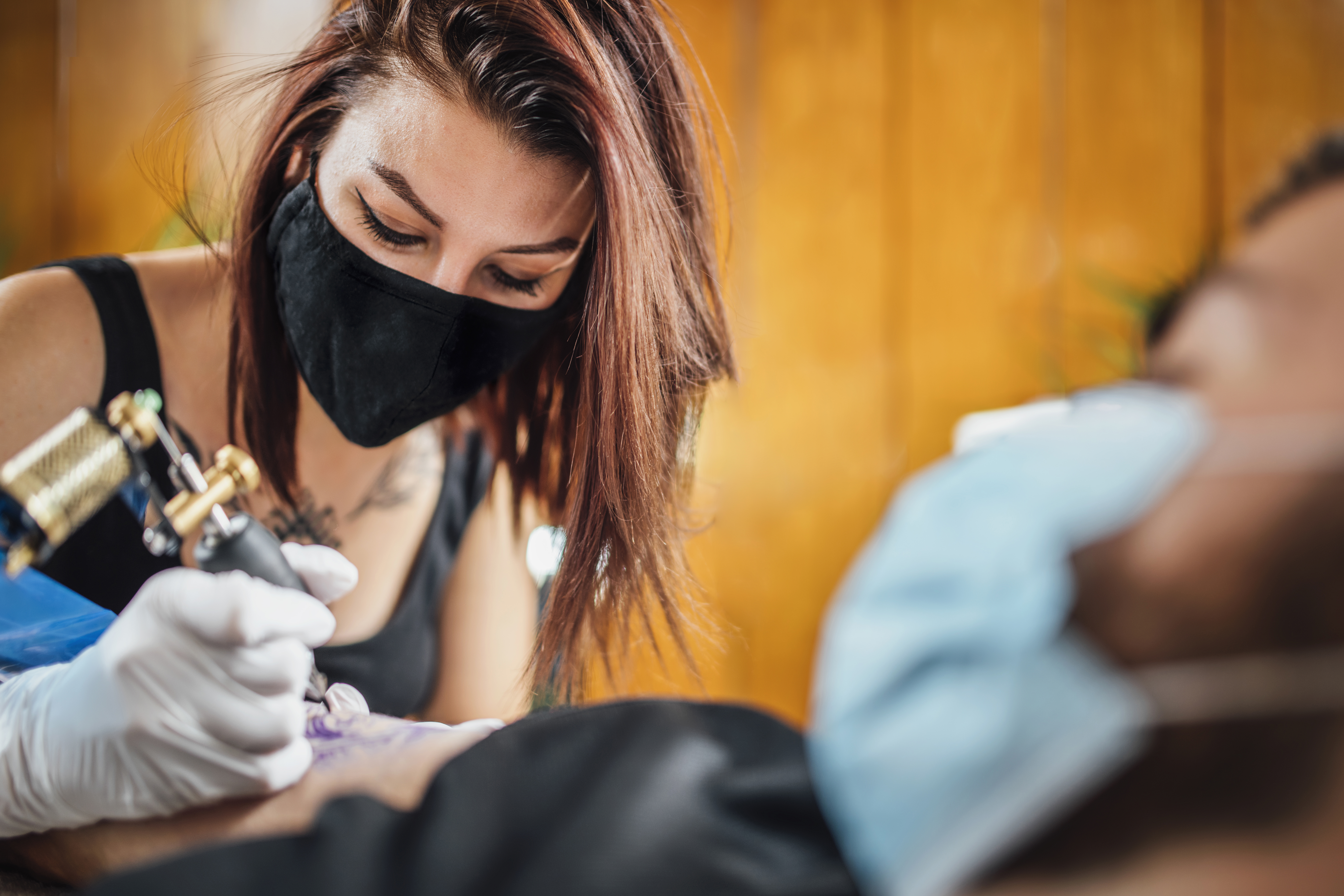 A masked woman tattoos a man’s arm.