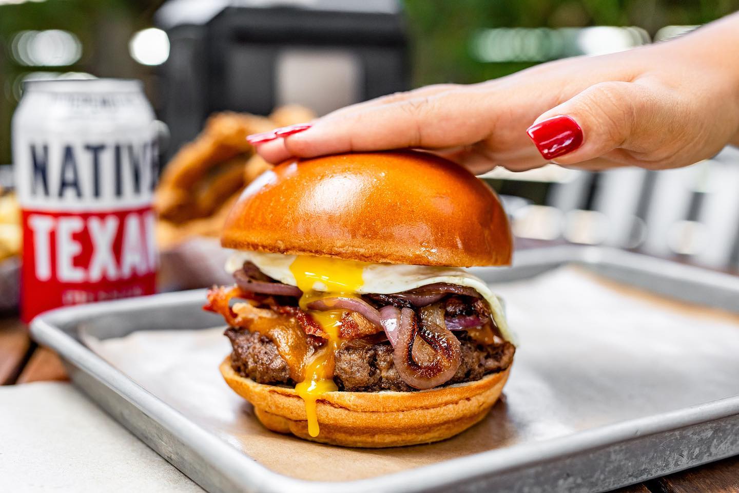 A manicured hand with long red nails resting on the top bun of a burger loaded with sauteed onions, bacon and a fried egg.