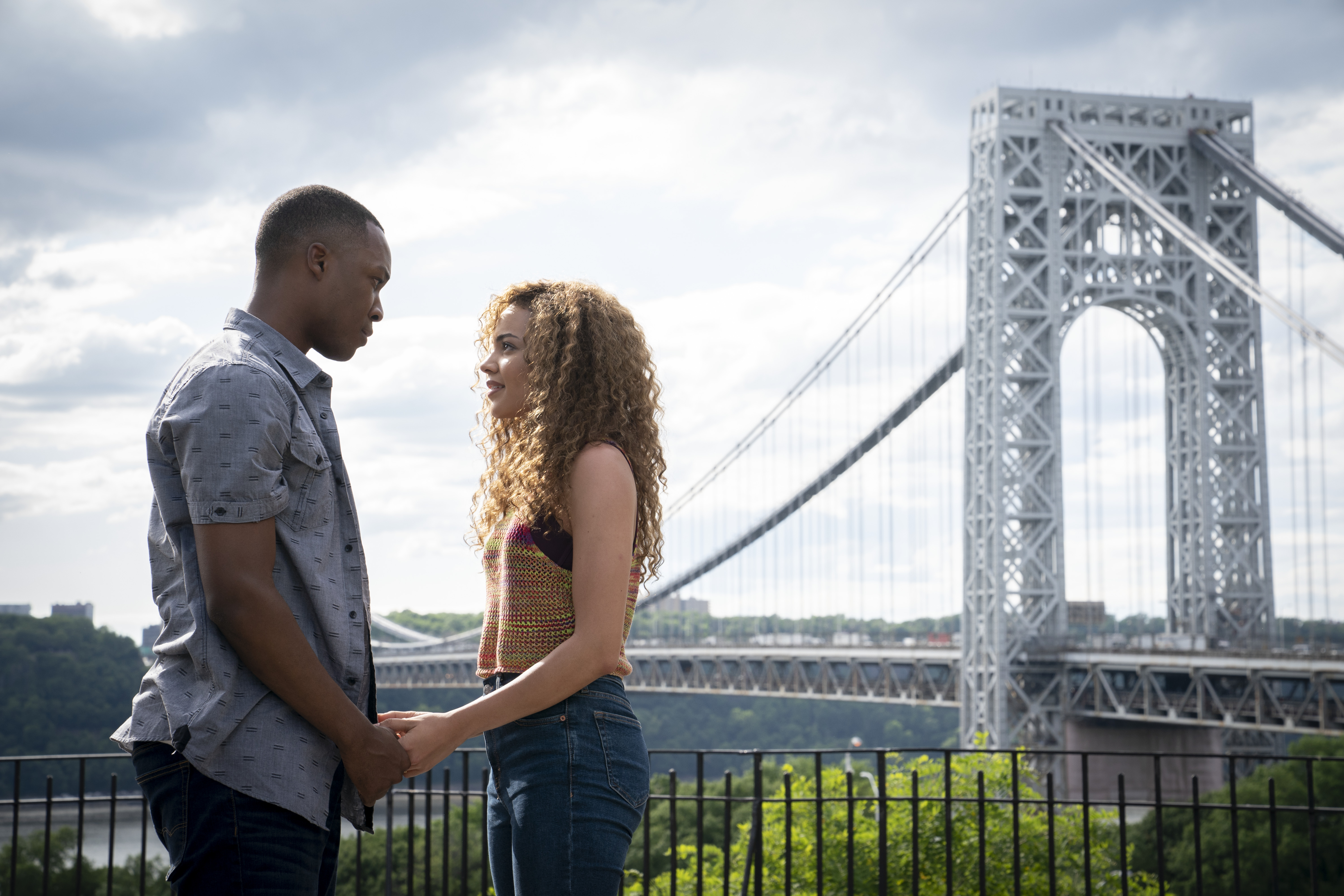 A couple stand facing one another while holding hands, with a New York bridge in the background.