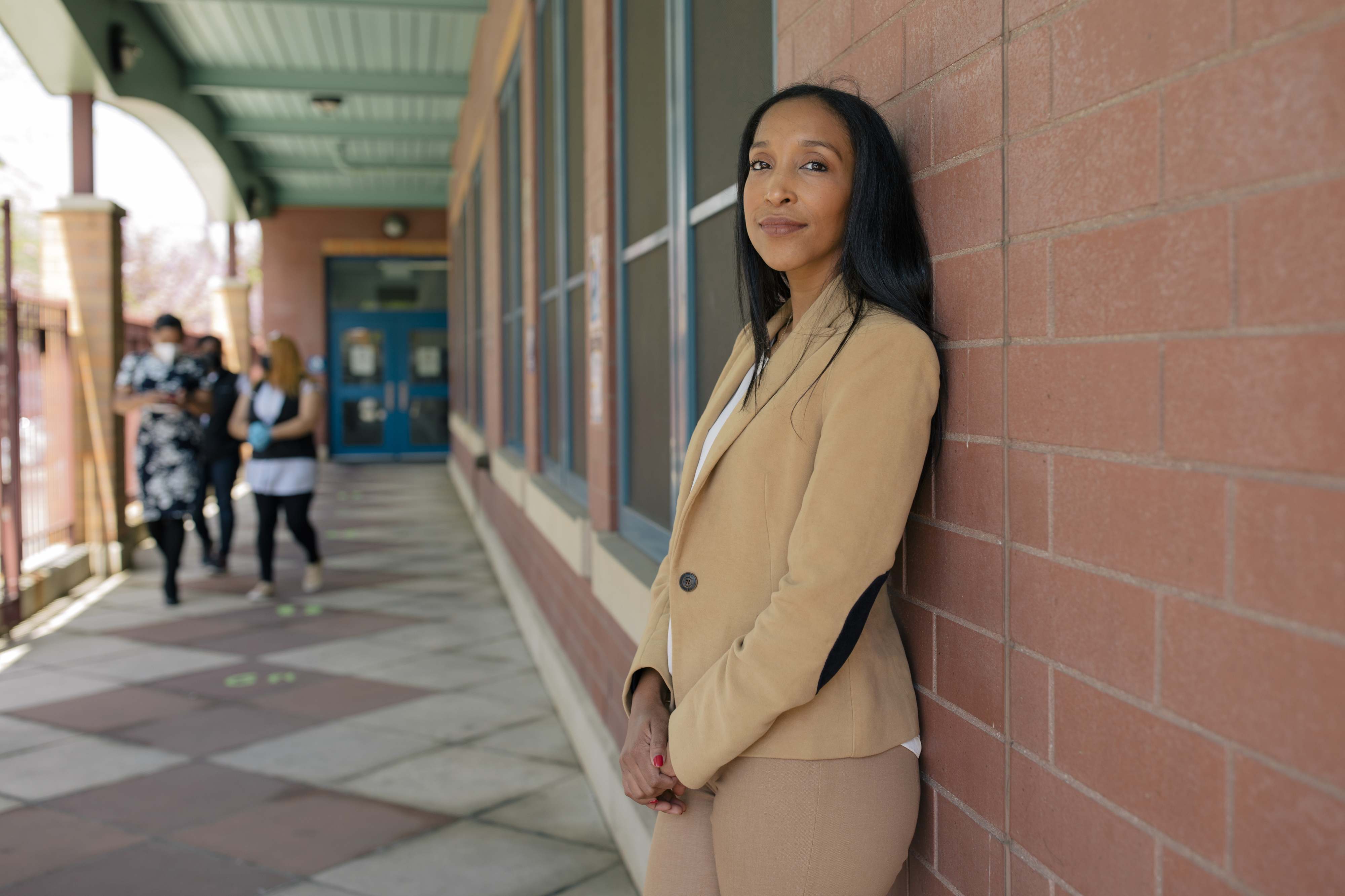 Yasirys Pichardo, wearing a tan pantsuit, stands against a red brick wall lined with windows while three women walk down the the path outside of P.S. 89.