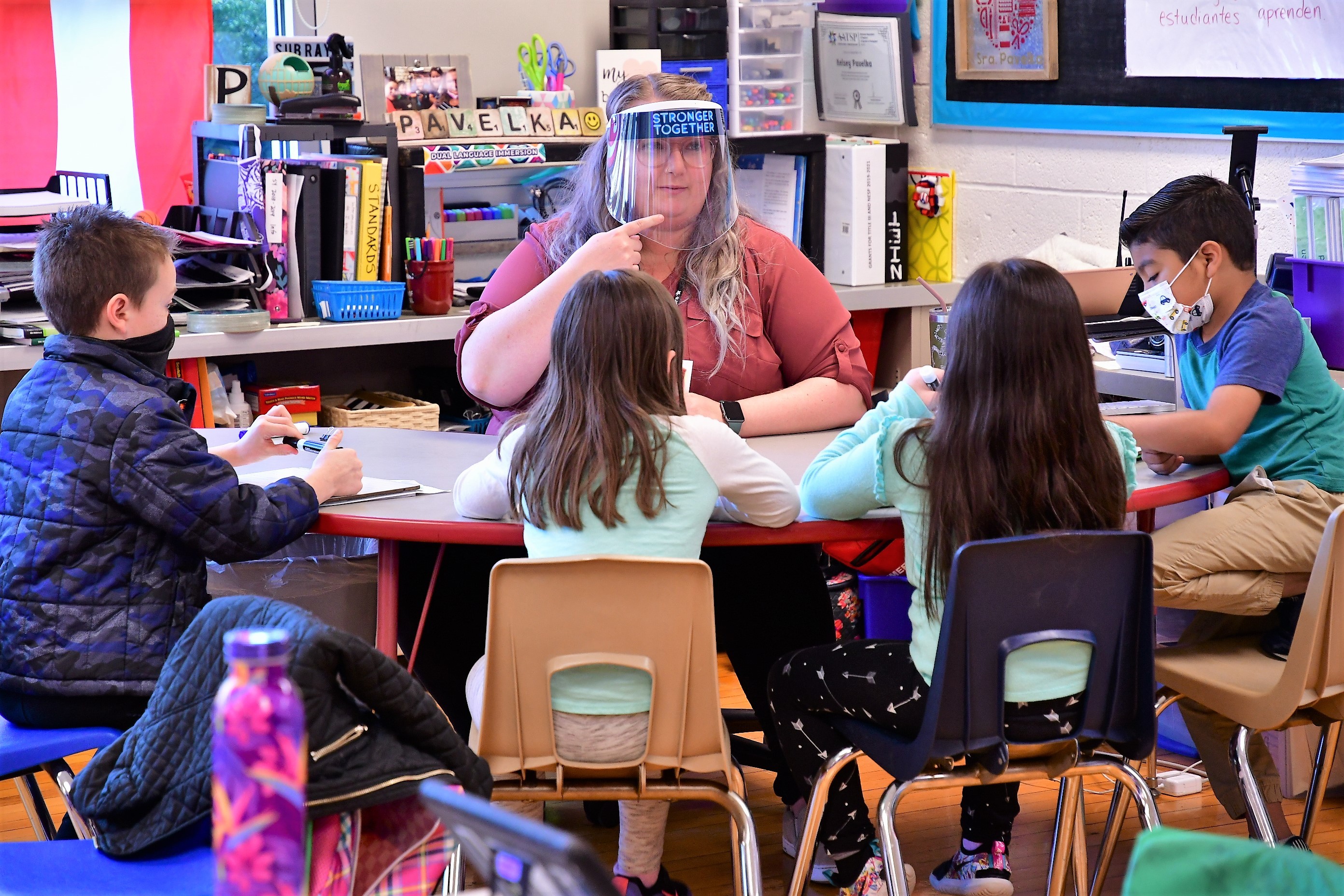 A teacher sits at a table facing four students. The teacher wears a clear plastic face shield and points to her chin.