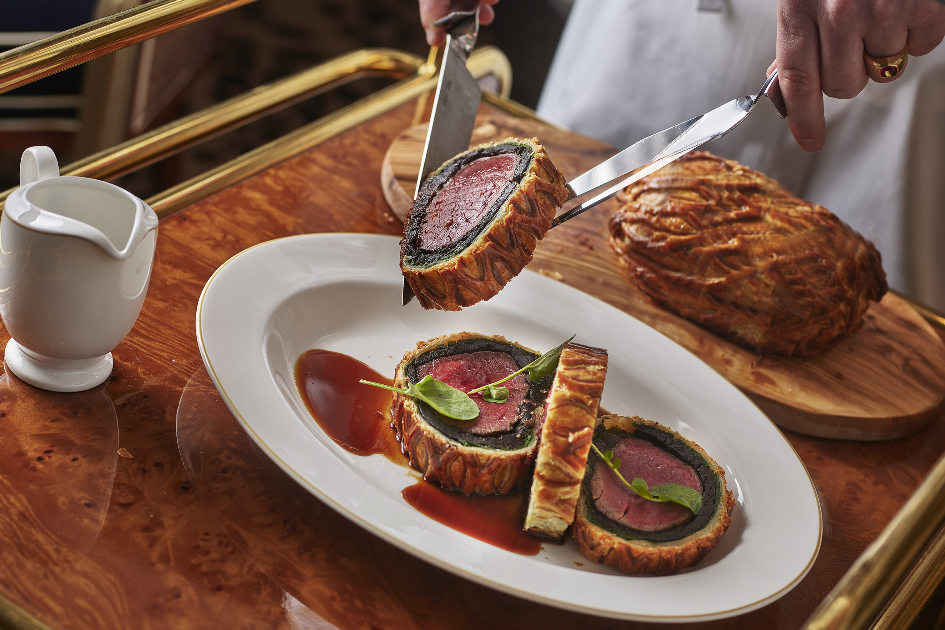 A sliced beef Wellington on an oval white plate with hands holding a slice in two knives