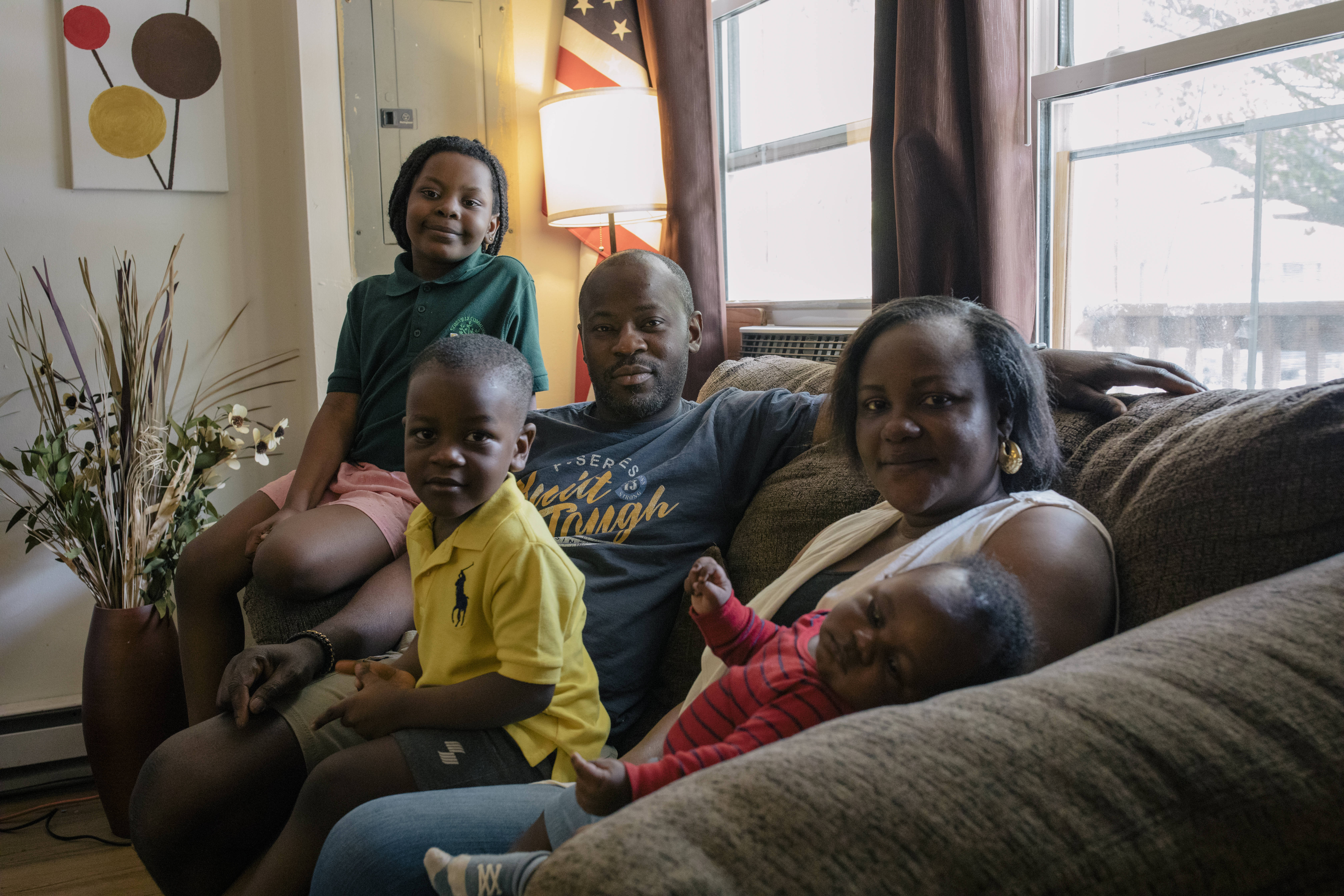 The Dairo family, consisting of parents John and Oyefunmilola and their young children, Abigail, Michael and infant Elijah, sit together on their living room couch next to a window.