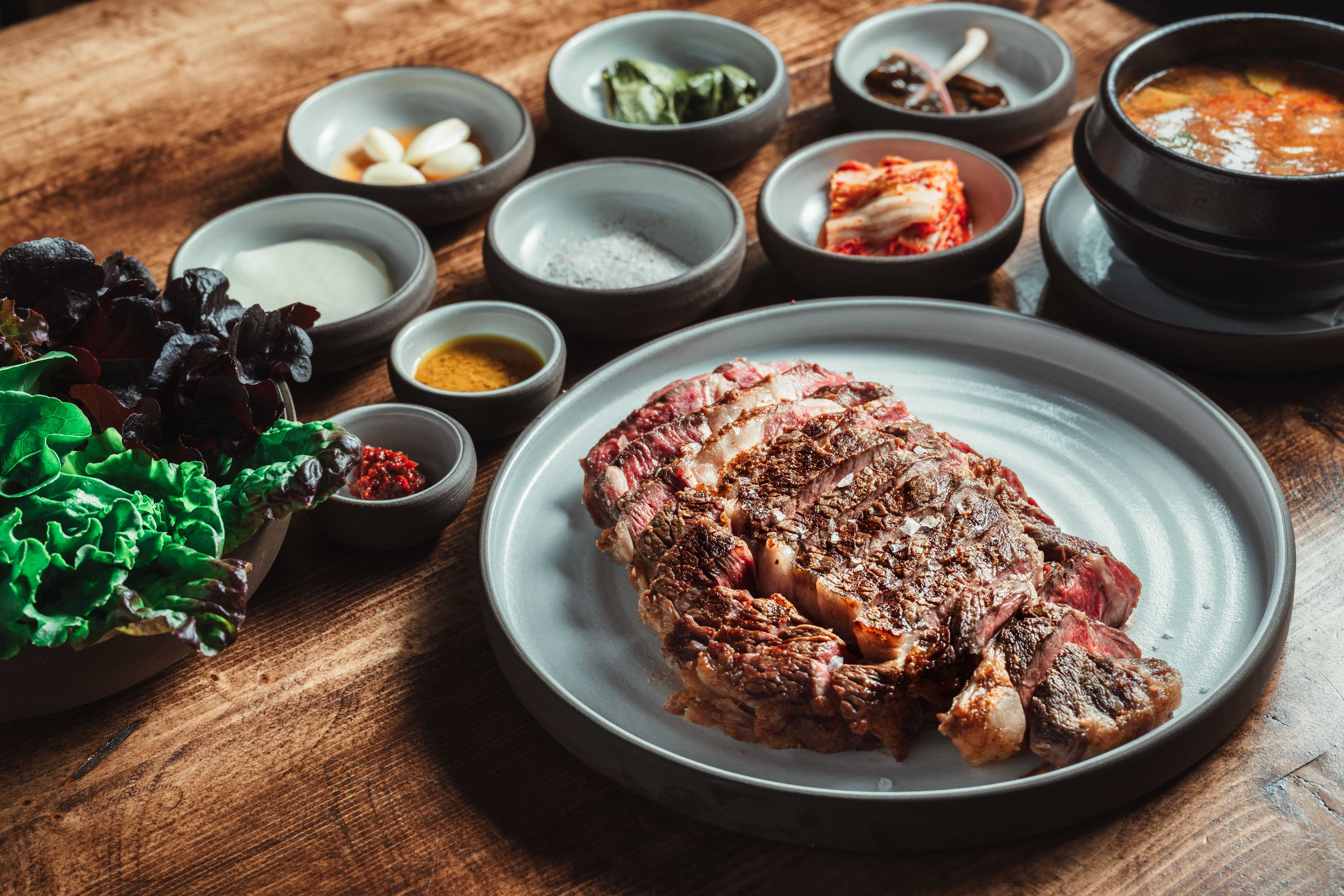 A sliced rare ribeye is on a gray plate on a wooden table. Small plates of Korean-style side dishes are visible in the background.
