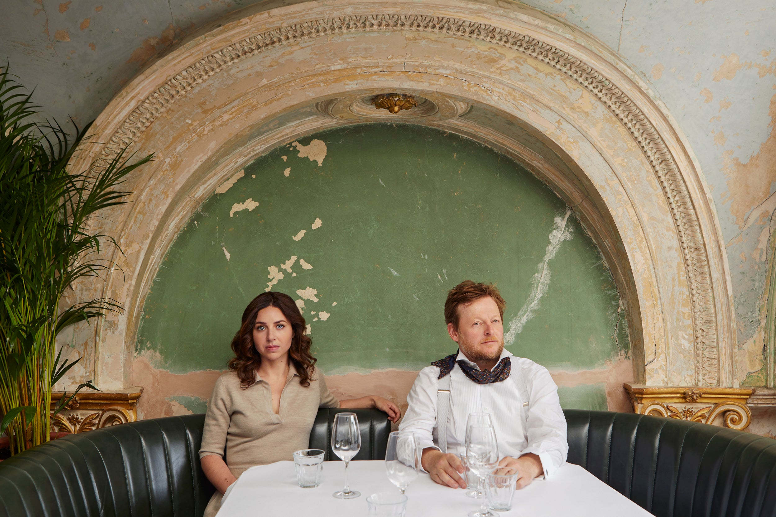 Florence Knight (left) and Jonny Gent (right) sit in a banquette with a white tablecloth and wine glasses, backed by a distressed green fresco with an arch
