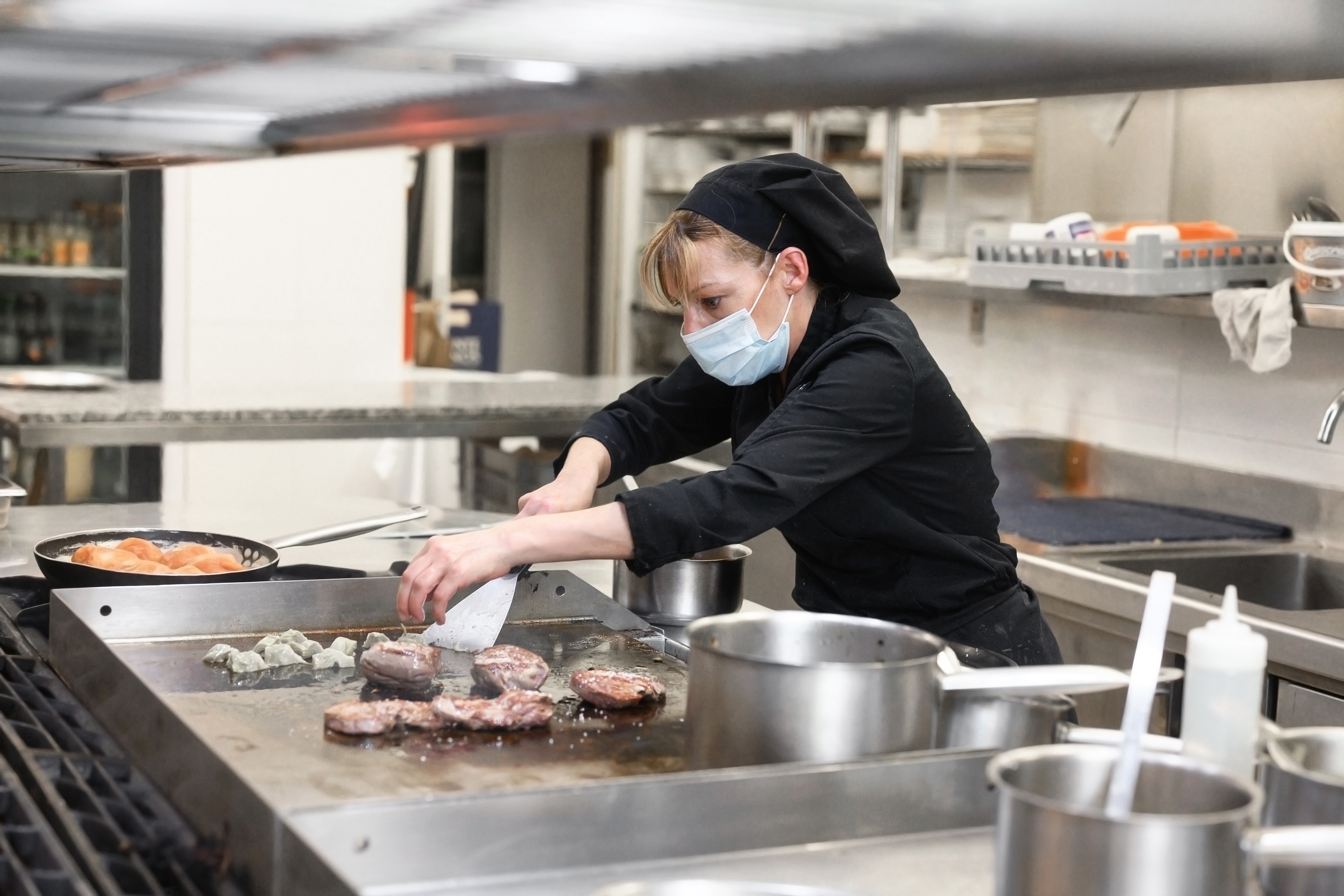 A female chef in a black chef’s coat and a surgical mask looks tense as she flips a steak on a griddle in a restaurant kitchen.