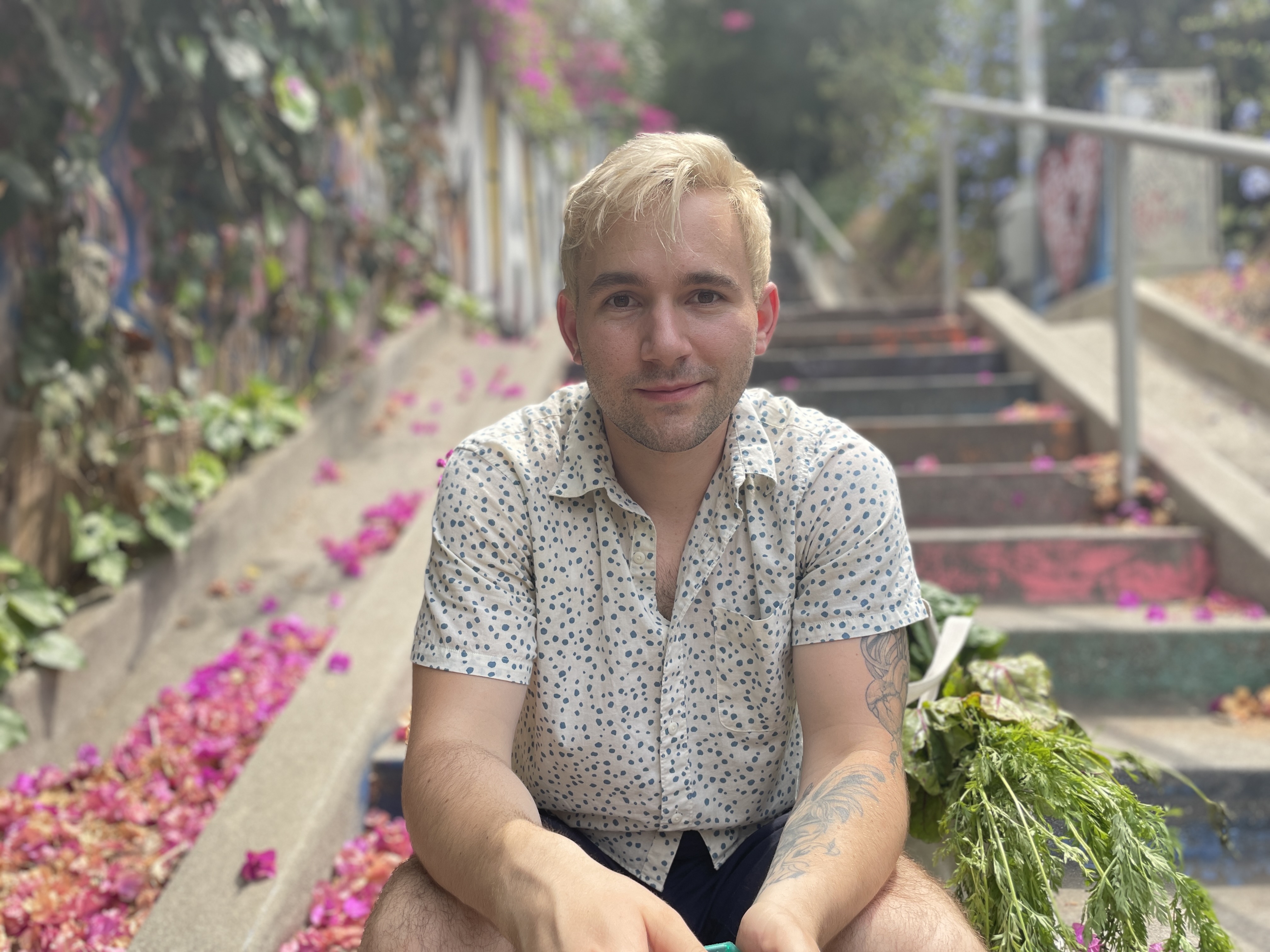 A white man in a short-sleeved blue and white collared shirt sits on cement steps with green ivy on the walls and pink petals on the ground in the background