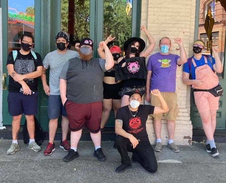 A group of seven employees wearing masks hold up fists outside the Voodoo Doughnut location in Portland’s Old Town.