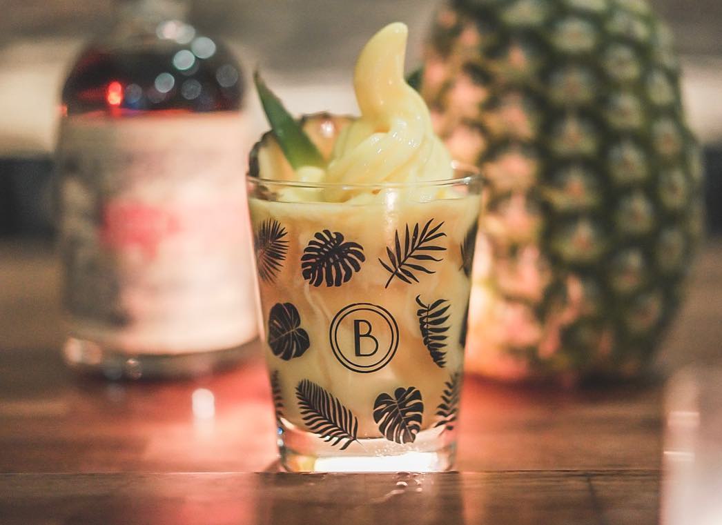 A boozy dole whip sits in a Blossom Bar branded glass, with a pineapple in the background