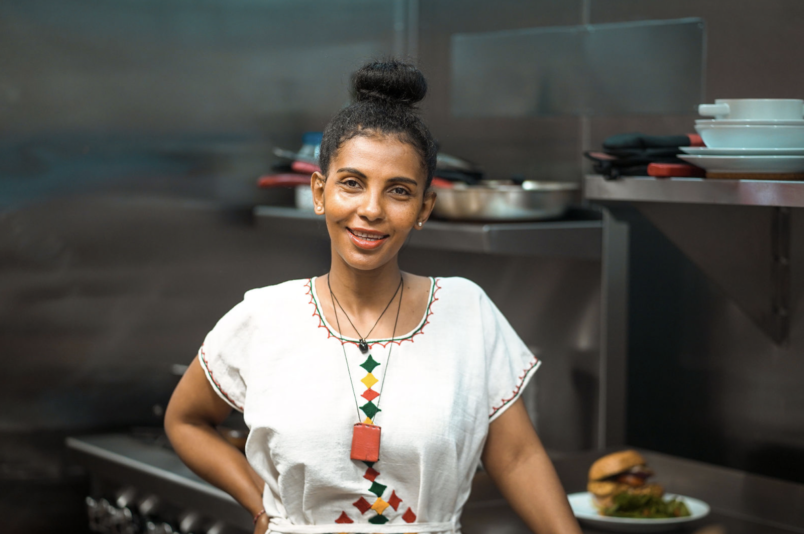 A chef in a white shirt smiles at the camera, hair up, inside of a kitchen.