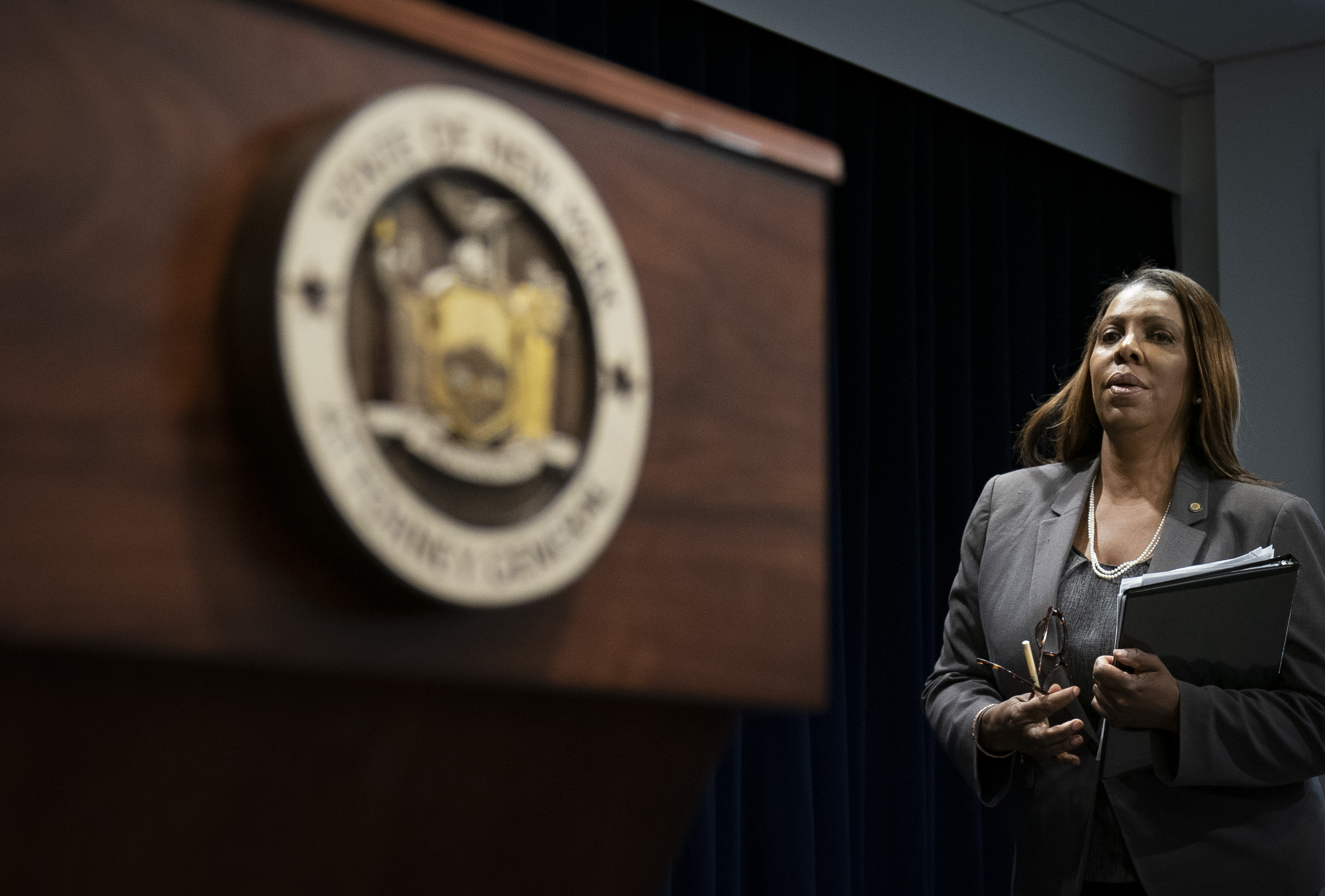 New York Attorney General Letitia James stands next to the state seal while holding a binder.