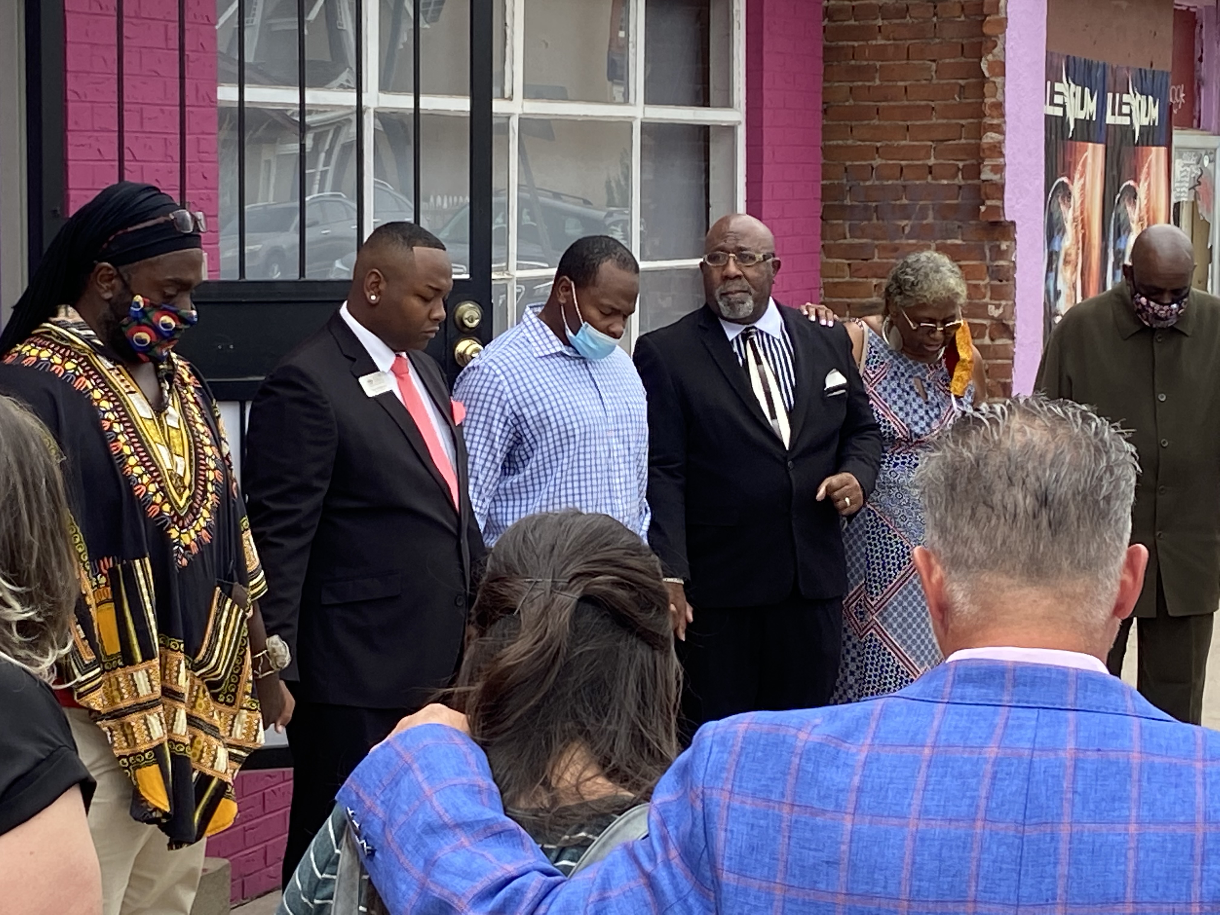 Denver school board member Tay Anderson, second from left, dressed in a suit and red tie, stands with his head bowed in prayer. He is standing in a circle of supporters holding hands.