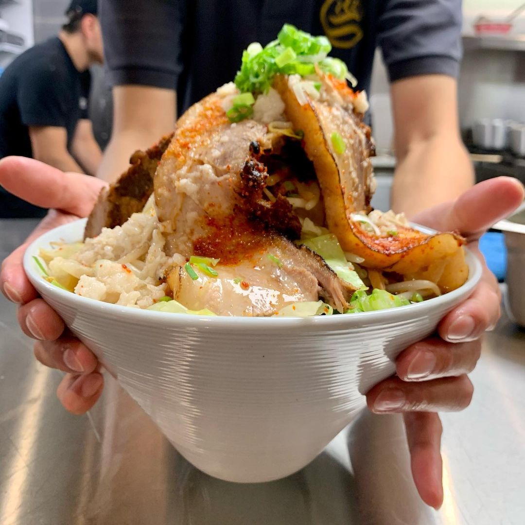Two hands hold a big white bowl full of ramen, with a mountain of pork belly and other toppings towering up over the top of the bowl