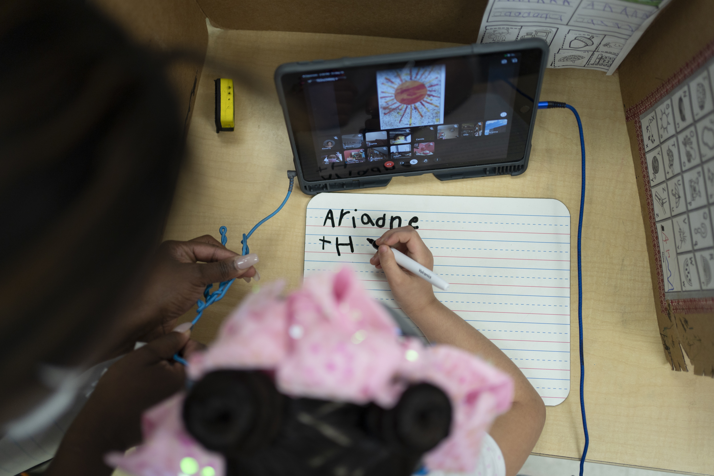 A girl wearing a pink bow works on a writing assignment while a teacher looks over her shoulder. There is a tablet on the desk in front of her.