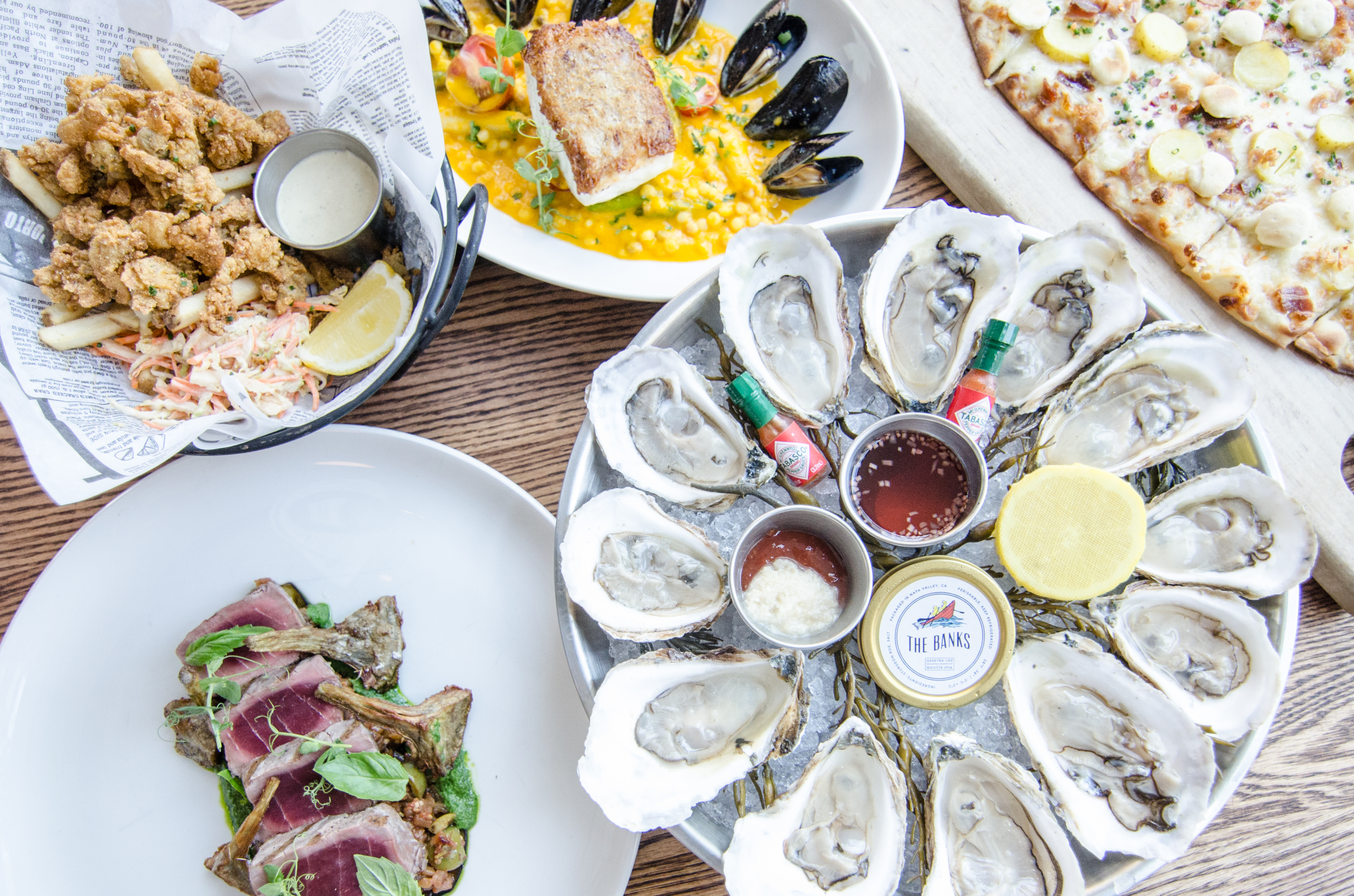 Overhead view of a table full of seafood dishes, including a platter of oysters, a basket of fried clams and fries, rare bluefin tuna, and more