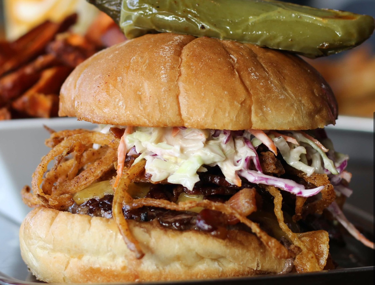 A large burger topped with frizzled onions, coleslaw, and a jalapeno pepper on top